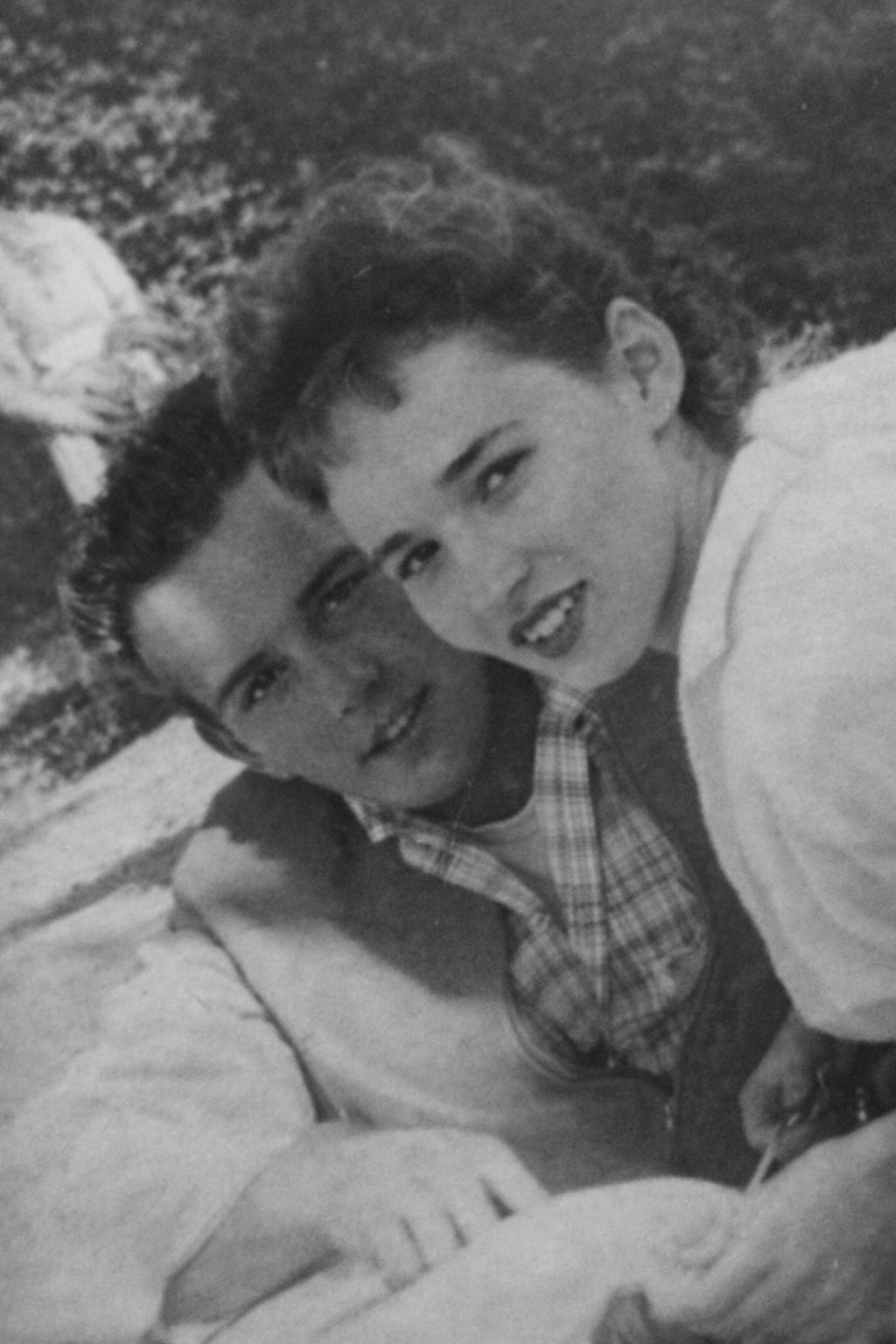 Black-and-white photo of a young man and woman smiling and close together outdoors, with grassy area in the background.