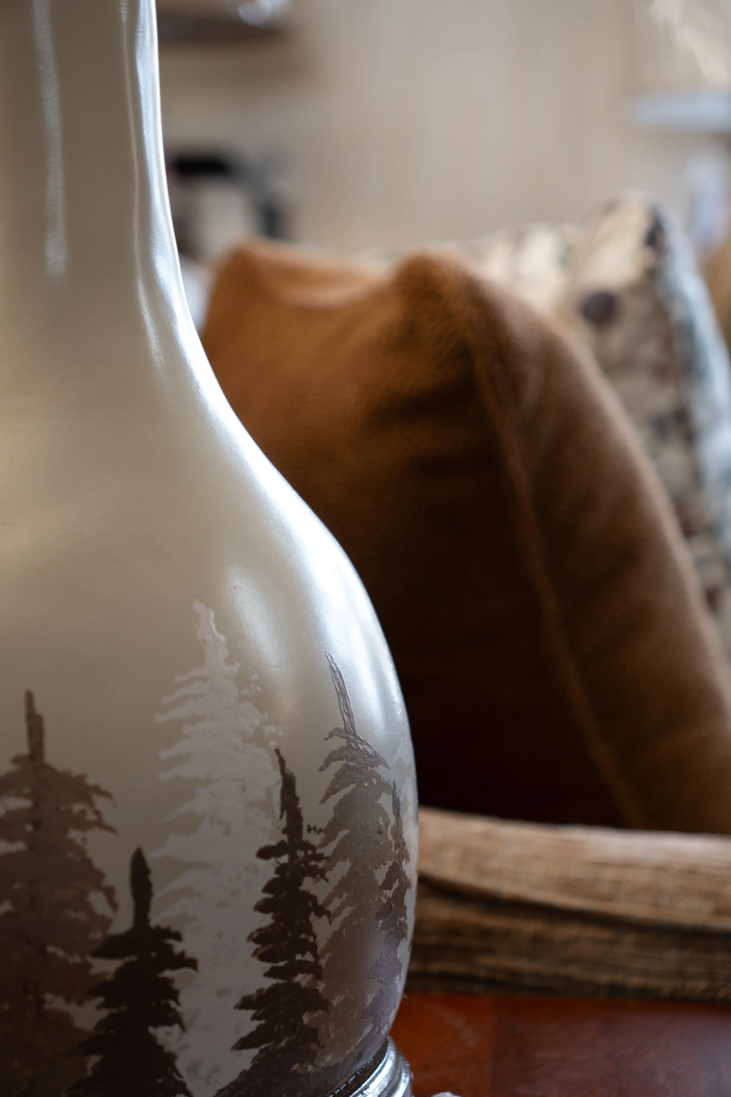 Close-up of a decorative vase with a painted landscape of pine trees, placed near a brown upholstered chair in a cozy indoor setting.