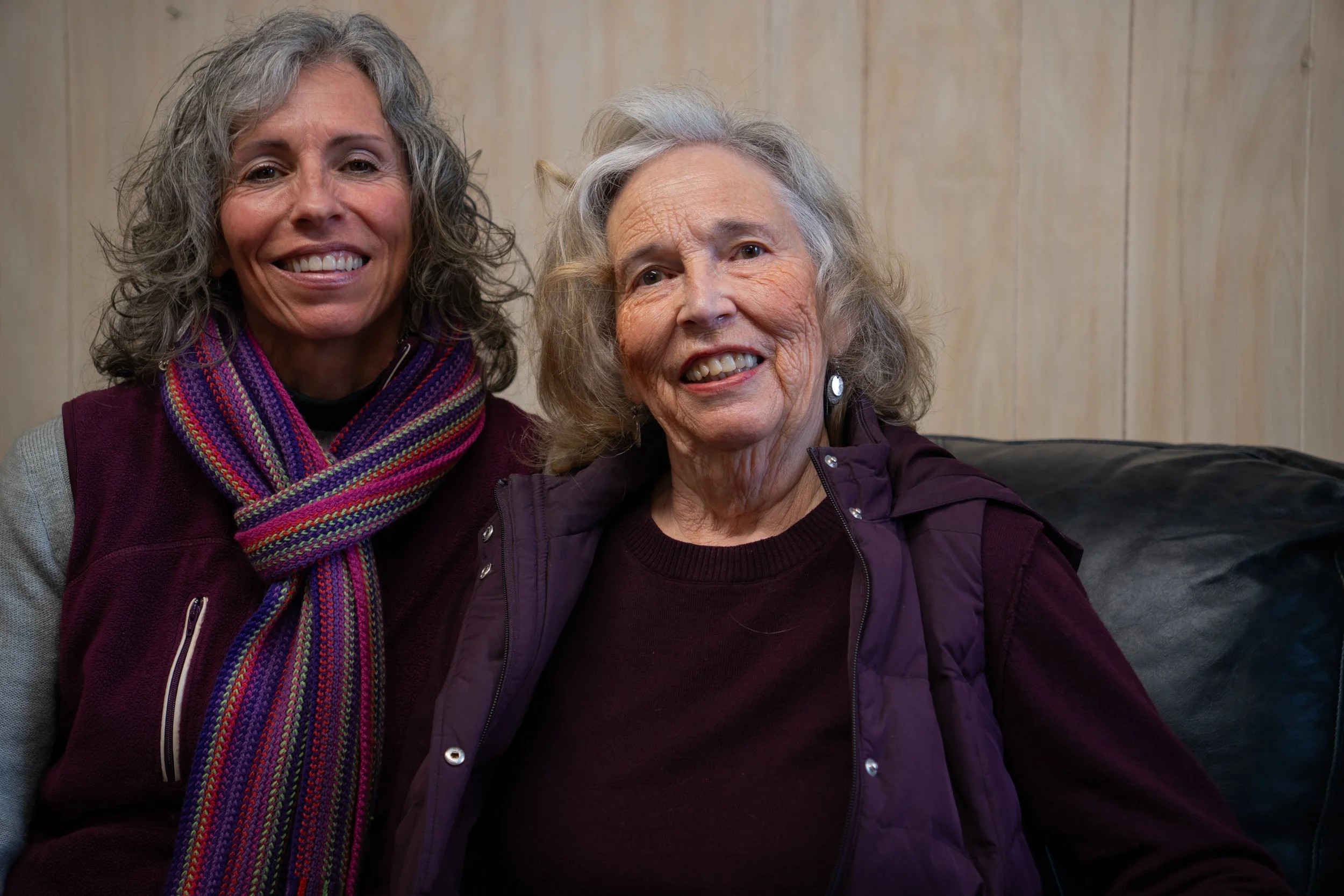 Two smiling women, one middle-aged and one elderly, sitting close together on a black sofa in front of a light-colored wooden wall, both wearing purple-toned clothing and smiling at the camera.
