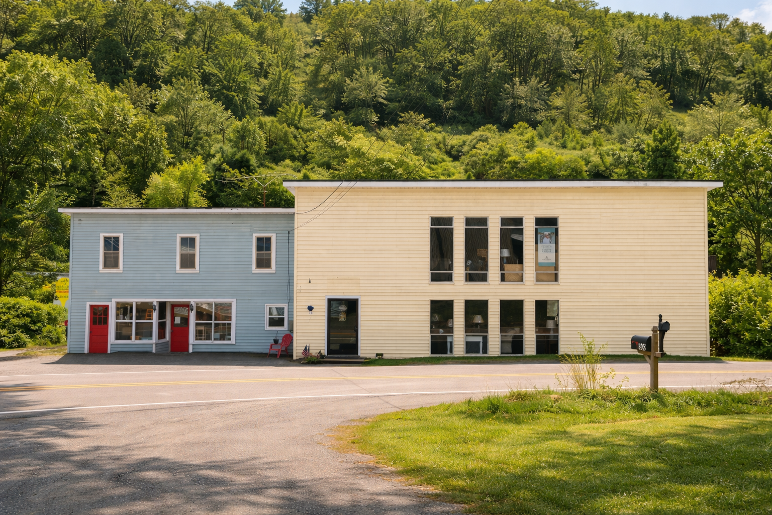A two-story building with a yellow and blue facade, red doors, and multiple windows, situated next to a road with a mailbox and green grass, with a forested hillside in the background.