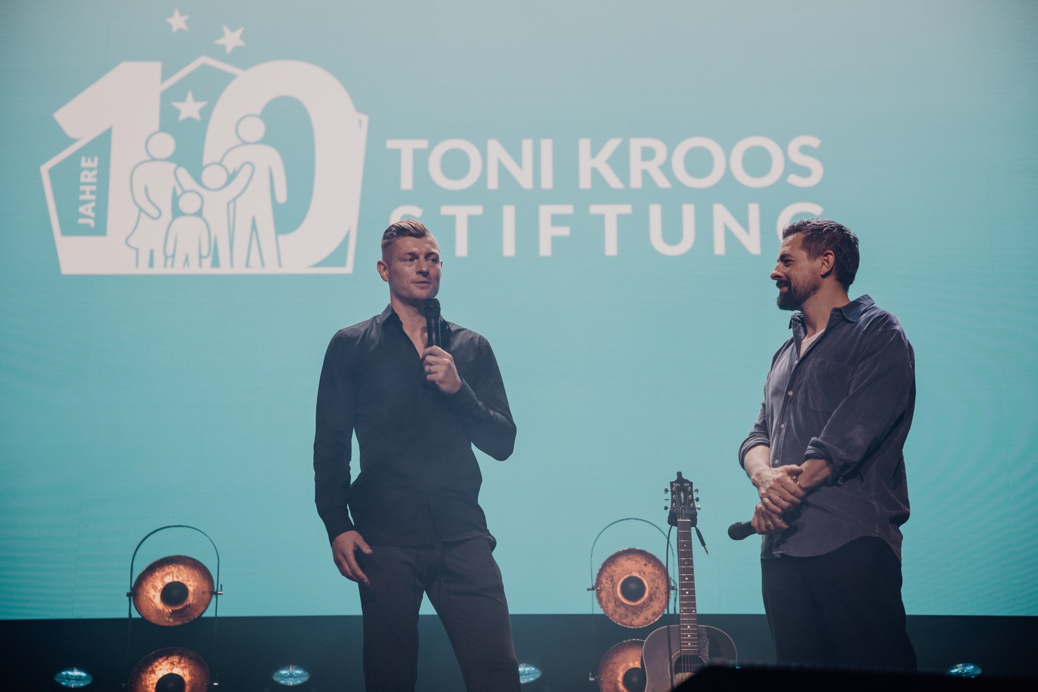 Zwei Männer auf einer Bühne bei einer Veranstaltung der Toni Kroos Stiftung. Einer spricht mit einem Mikrofon, im Hintergrund ein großes Logo der Stiftung und ein Gitarrenset.