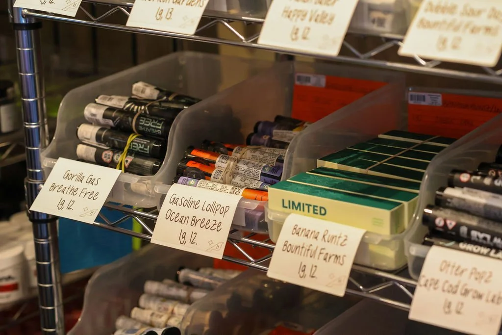 Storage shelf with containers of vape cartridges and e-liquids, and handwritten labels indicating different flavors and brands.