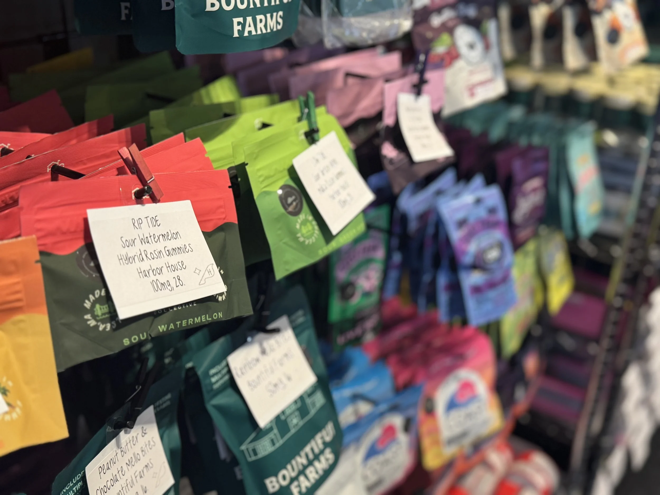 Colorful bags of candy or gummy toys hanging on a display rack at a store, with handwritten labels showing the flavor and brand.