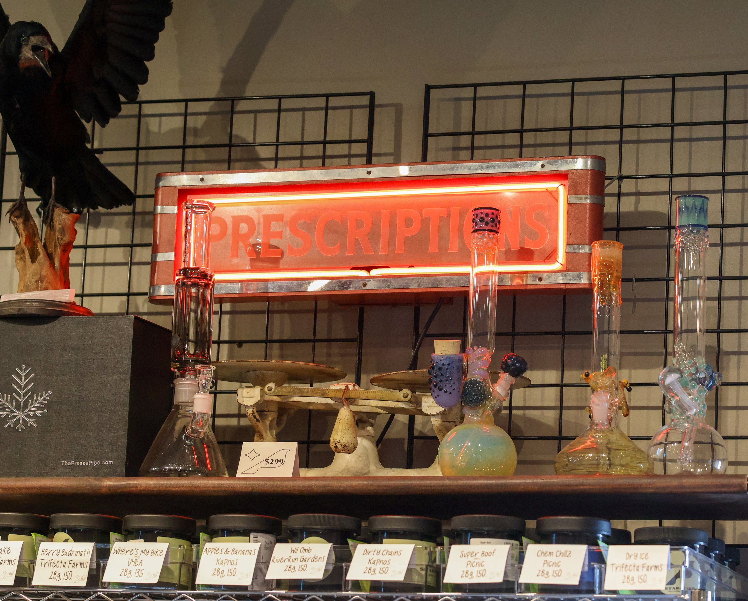 Display of glass smoking pipes and glassware on a store shelf with a neon 'Prescriptions' sign above. Various glass pieces are arranged on the shelf, and labels with prices are in front of the jars below.