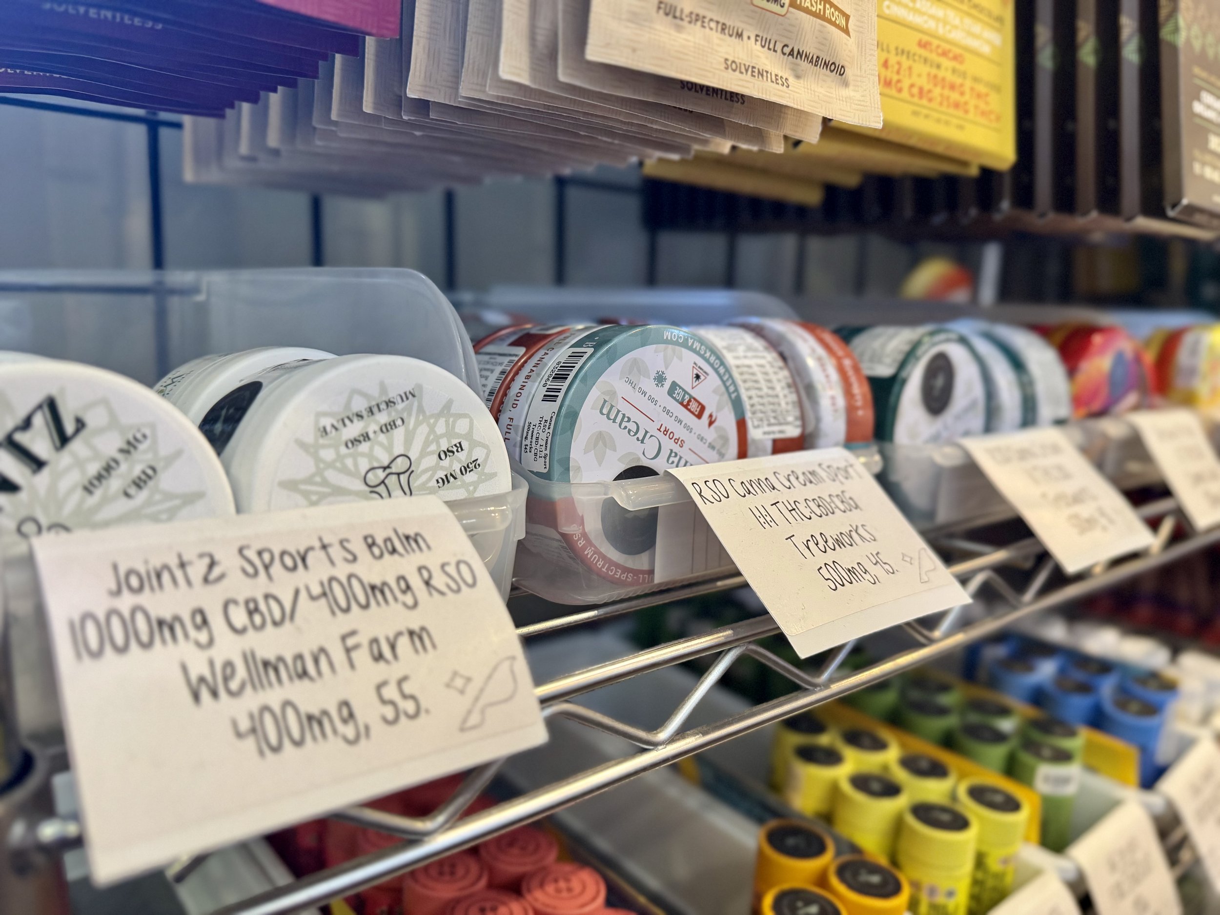 Shelf of CBD products, including jars of topical cream and labeled notes with product and dosage information in a store.