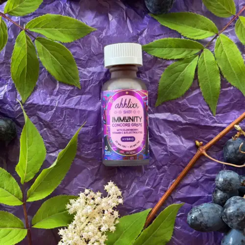 A small bottle of immuninity supplement with a purple label, placed on purple crinkled paper, surrounded by green leaves, white flowers, and a cluster of dark grapes.