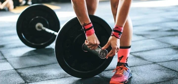 Person lifting a barbell with weights at a gym, wearing bright pink socks and athletic shoes.