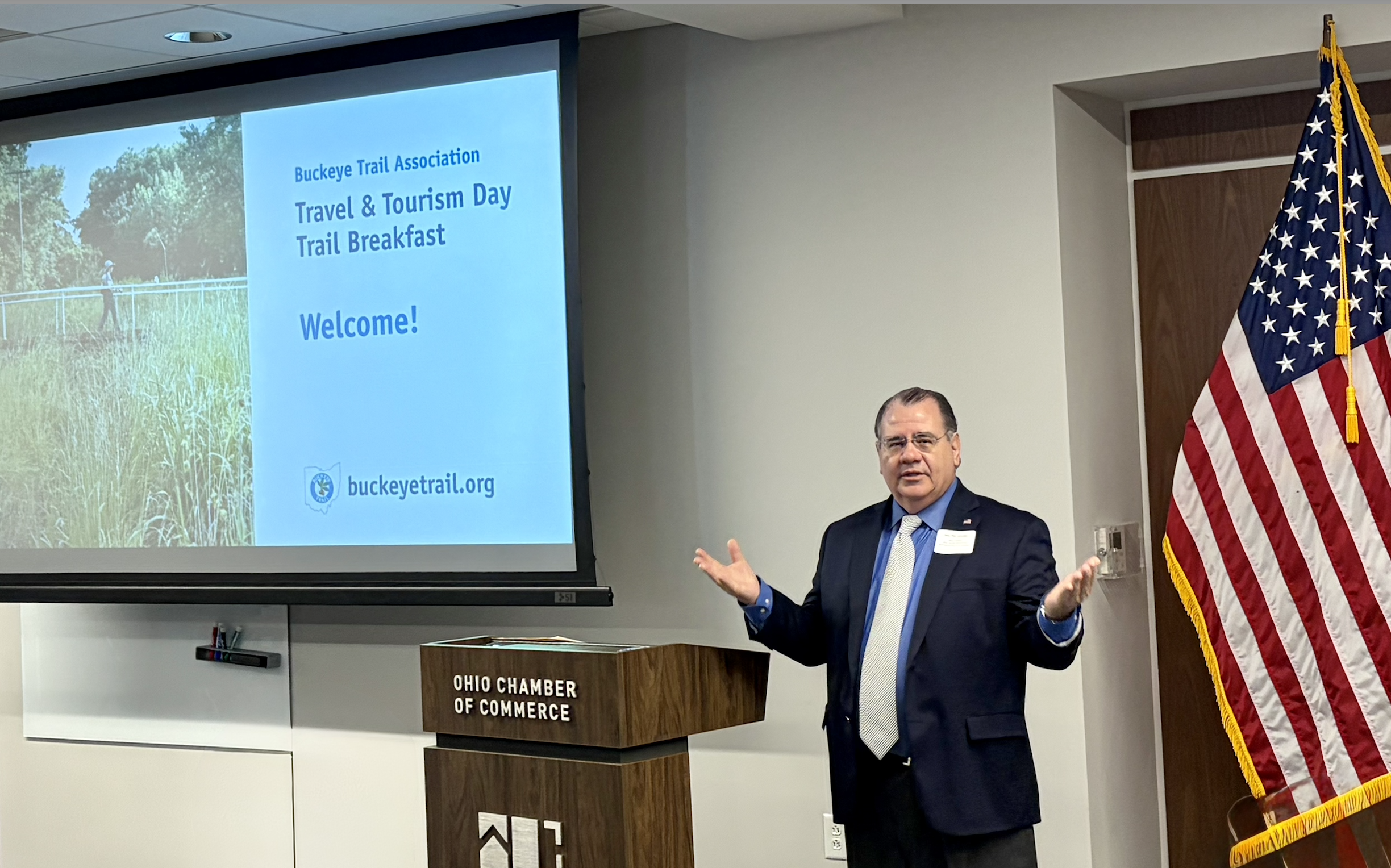 Sen. Tim Schaffer speaks at the Trail Breakfast, standing beside a podium with a screen reading Travel & Tourism Day Trail Breakfast.