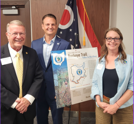 Sen. Steve Wilson, the Director of Travel and Tourism Policy at the Ohio Chamber of Commerce, and Jill Moorhead stand together holding a framed Buckeye Trail map.