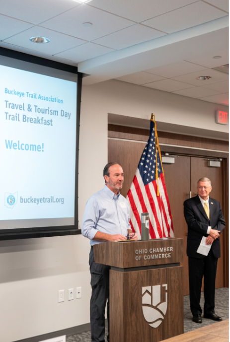 Buckeye Trail Association Executive Director Andrew Bashaw and Sen. Steve Wilson stand near a podium in front of a screen reading Travel & Tourism Day Trail Breakfast.