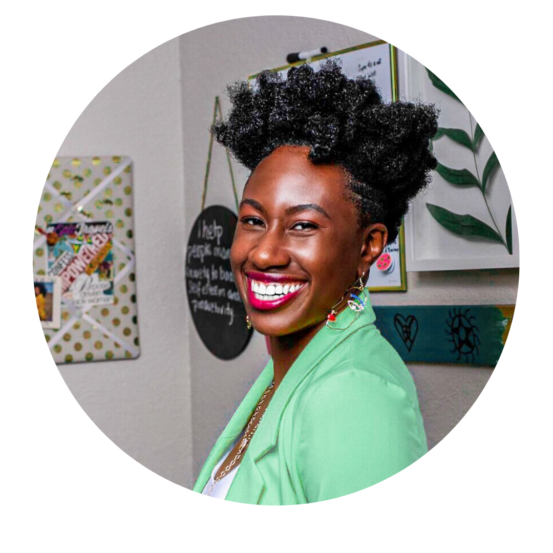 A smiling woman with natural curly hair styled in an updo, wearing a green blazer and colorful earrings, standing in an office or classroom setting with bulletin boards and decorations in the background.