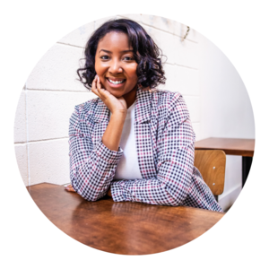 A young woman with curly hair smiling, sitting at a wooden table indoors, wearing a checkered blazer.