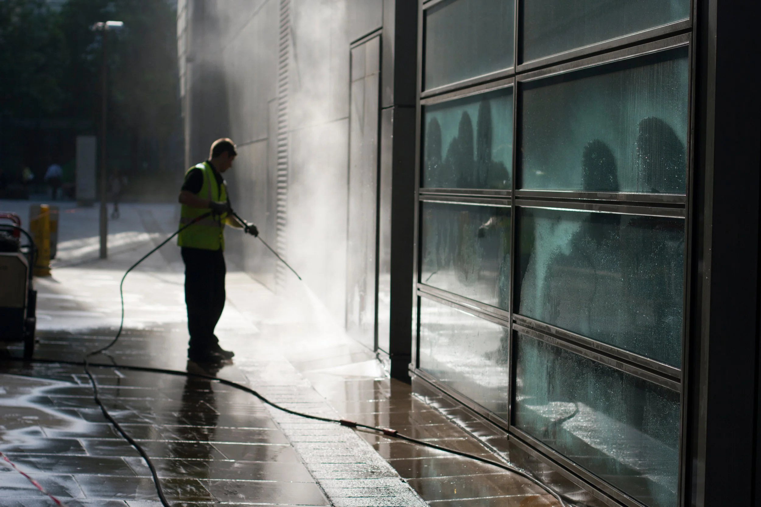 Worker in a yellow safety vest using a pressure washer to clean the exterior glass panels of a building, with sunlight and shadows visible.