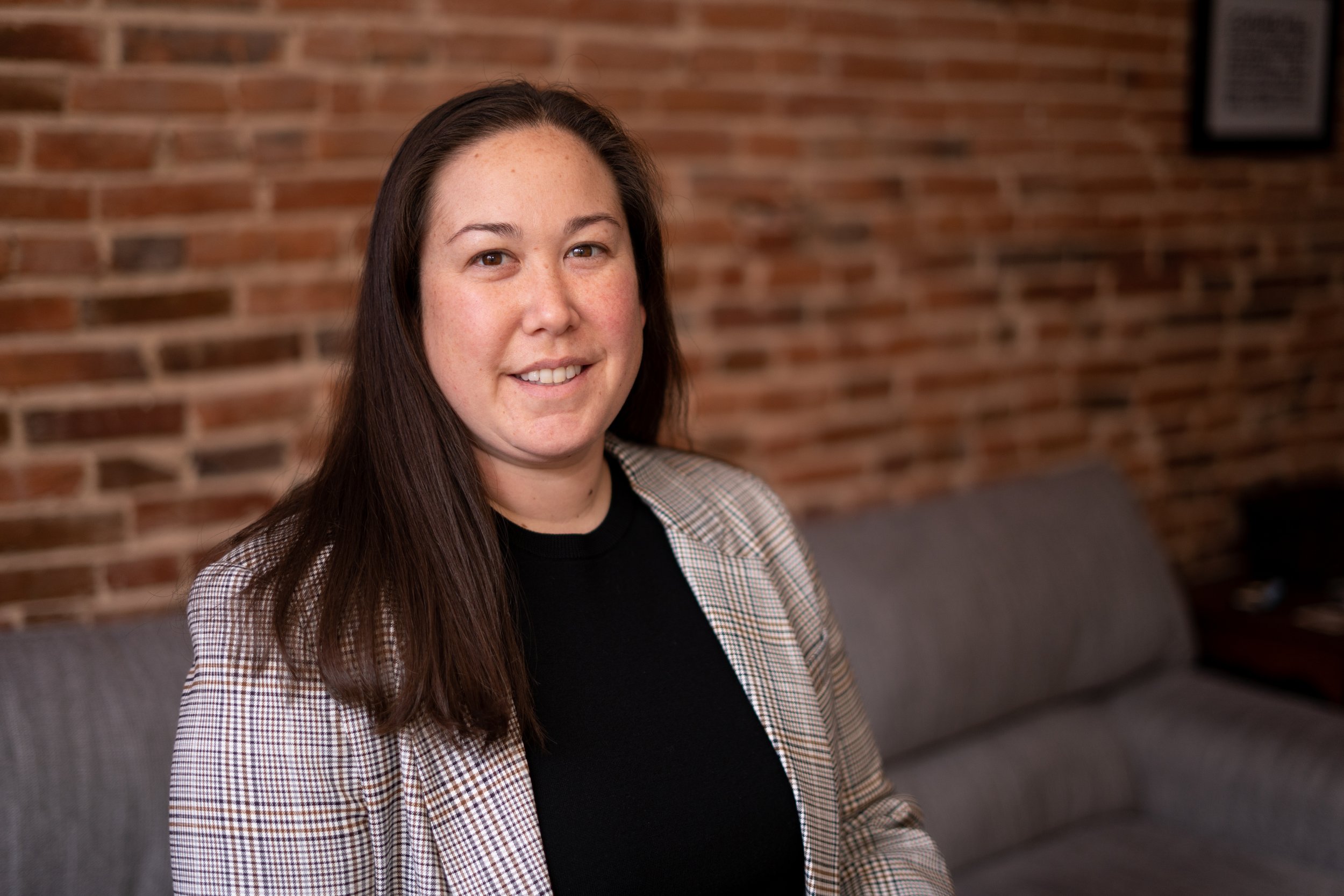 A woman with long brown hair, wearing a plaid blazer and black top, smiling while sitting in a room with a brick wall background.