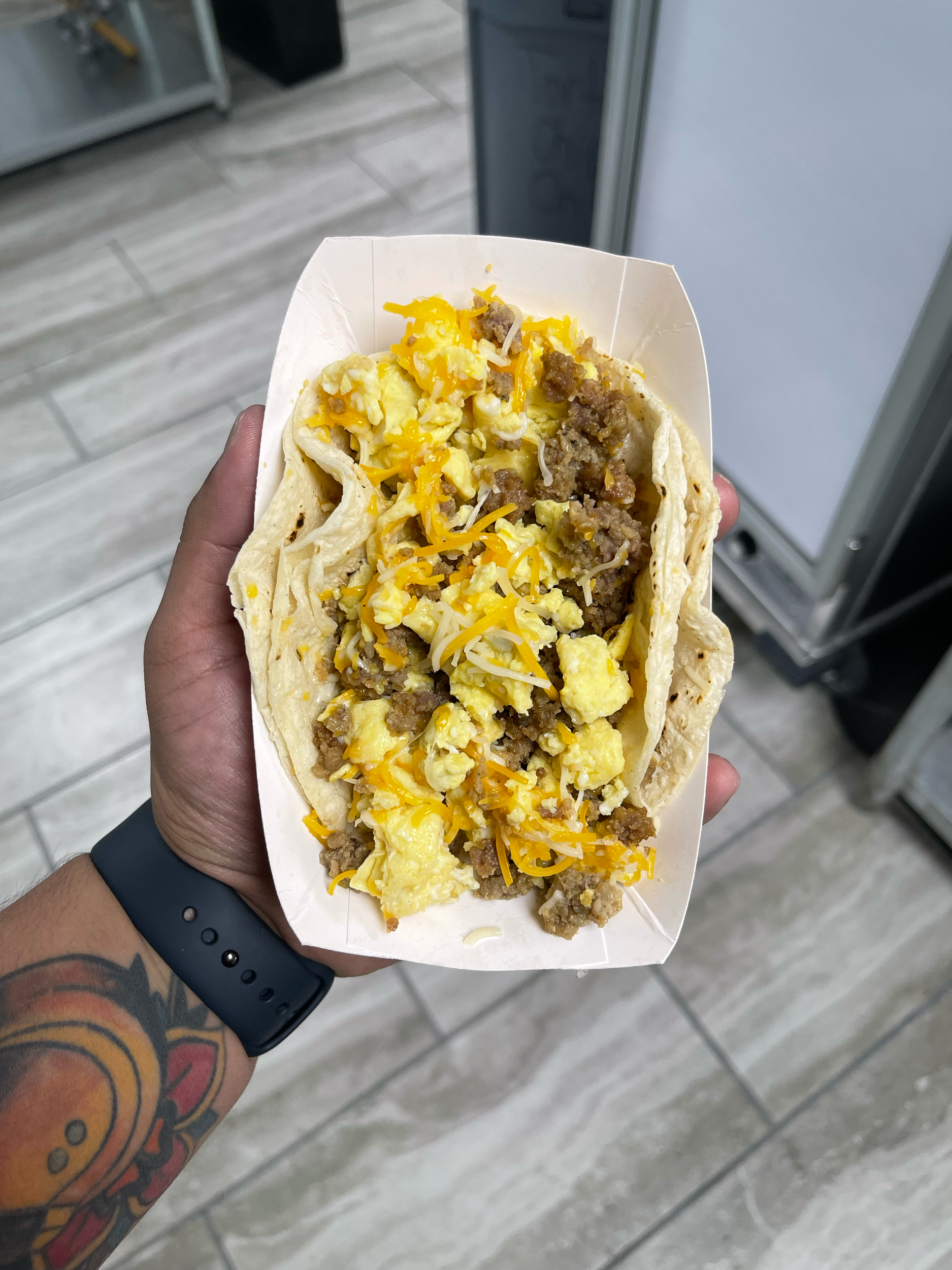 A hand holding a breakfast taco filled with scrambled eggs, ground beef, and shredded cheese. The person is wearing a black wristband and has a colorful tattoo on their arm. The background shows a tiled floor and part of a metal appliance.