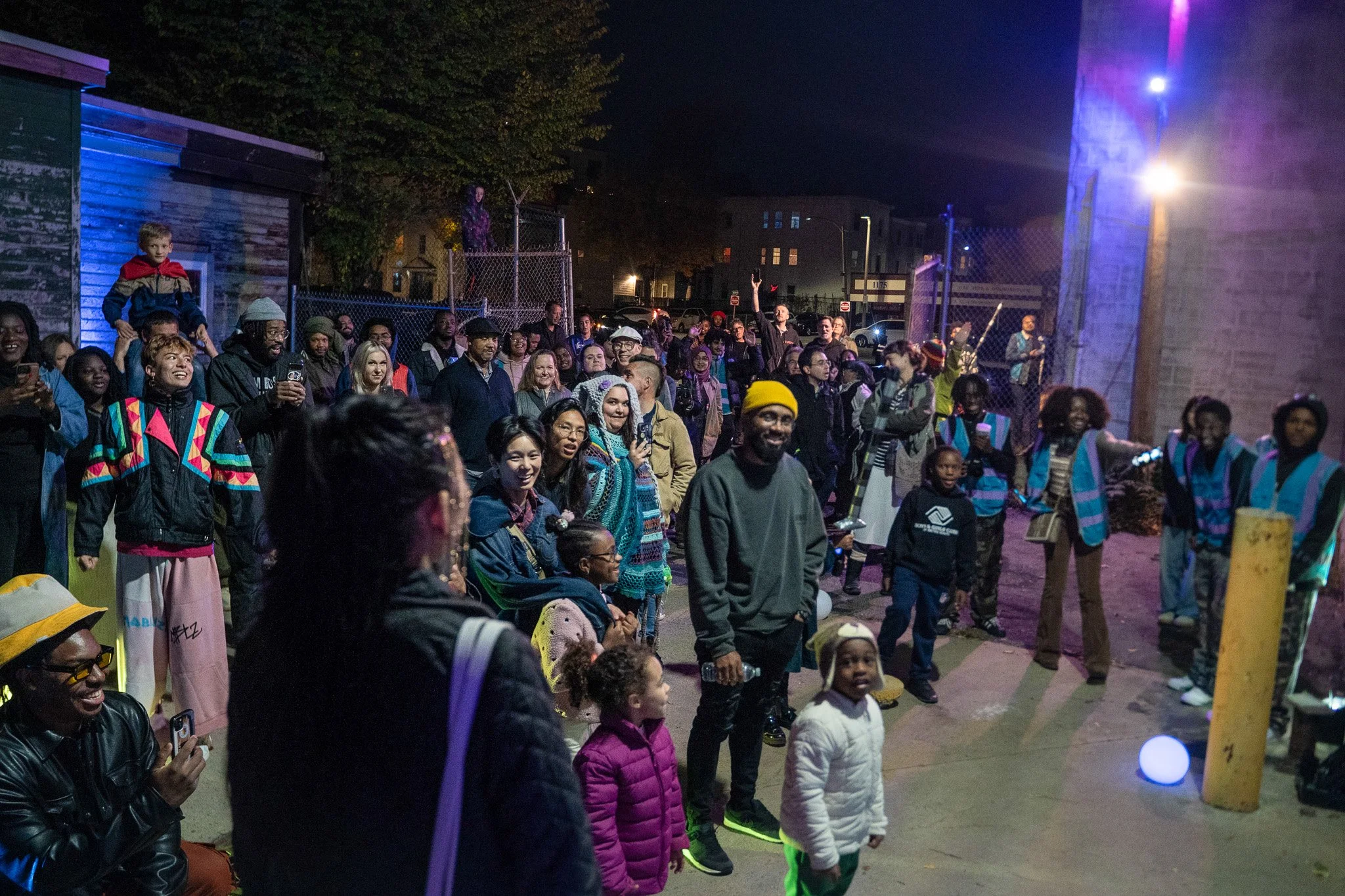 A diverse group of people, including children and adults, gathered outdoors at night, watching and enjoying a spotlight or performance near a brick wall and chain-link fence with colorful lights illuminating the scene.