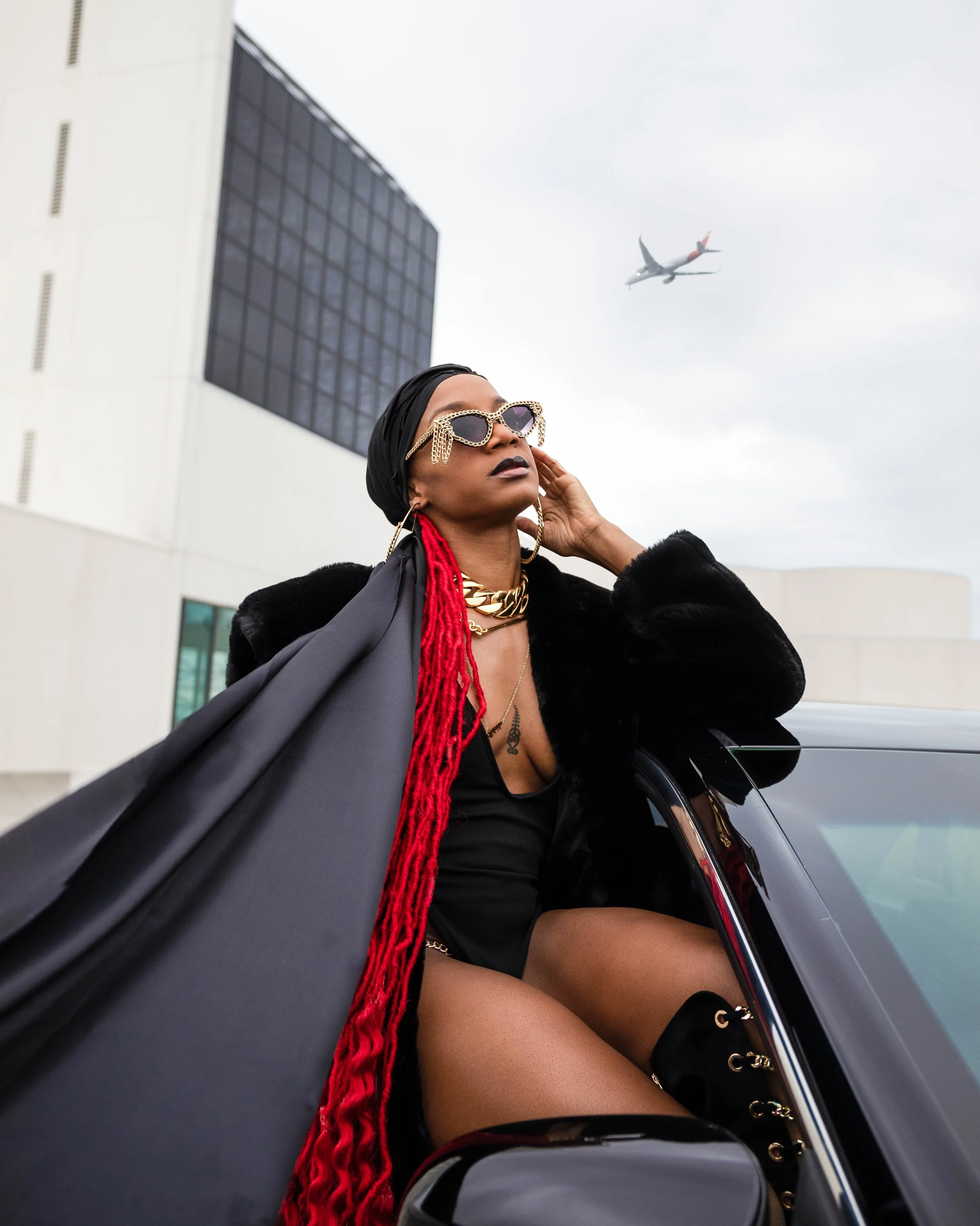 A woman with luxurious accessories looking confident while sitting in a black car against an urban backdrop with a plane flying overhead.