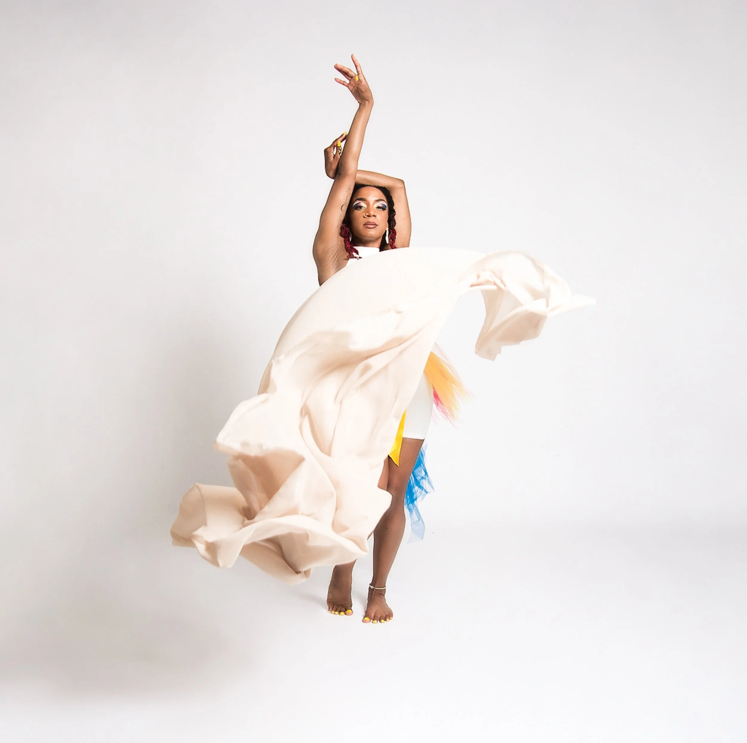 A woman dancing with one arm raised and the other behind her head, wearing a flowing beige skirt and colorful accessories, standing against a plain white background.