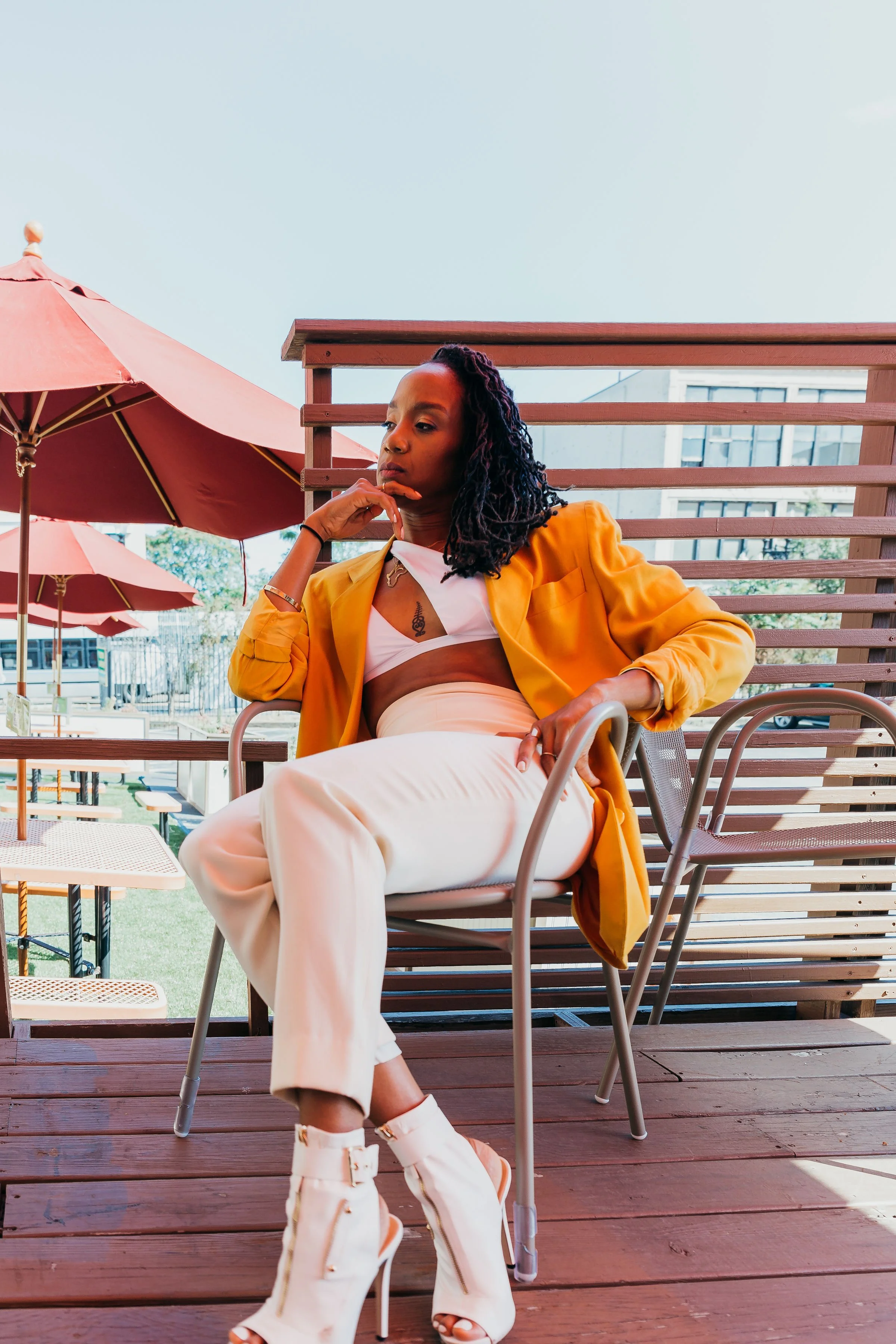 A woman sitting on a wooden patio chair, wearing a yellow blazer, white pants, and high-heeled boots, with a yellow umbrella and outdoor furniture in the background.