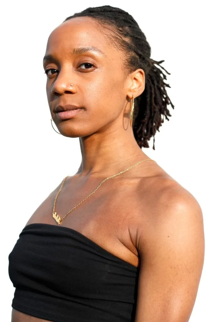 Portrait of a woman with short microlocs, wearing a black strapless top, gold hoop earrings, a nose stud, and a gold necklace, against a white background.