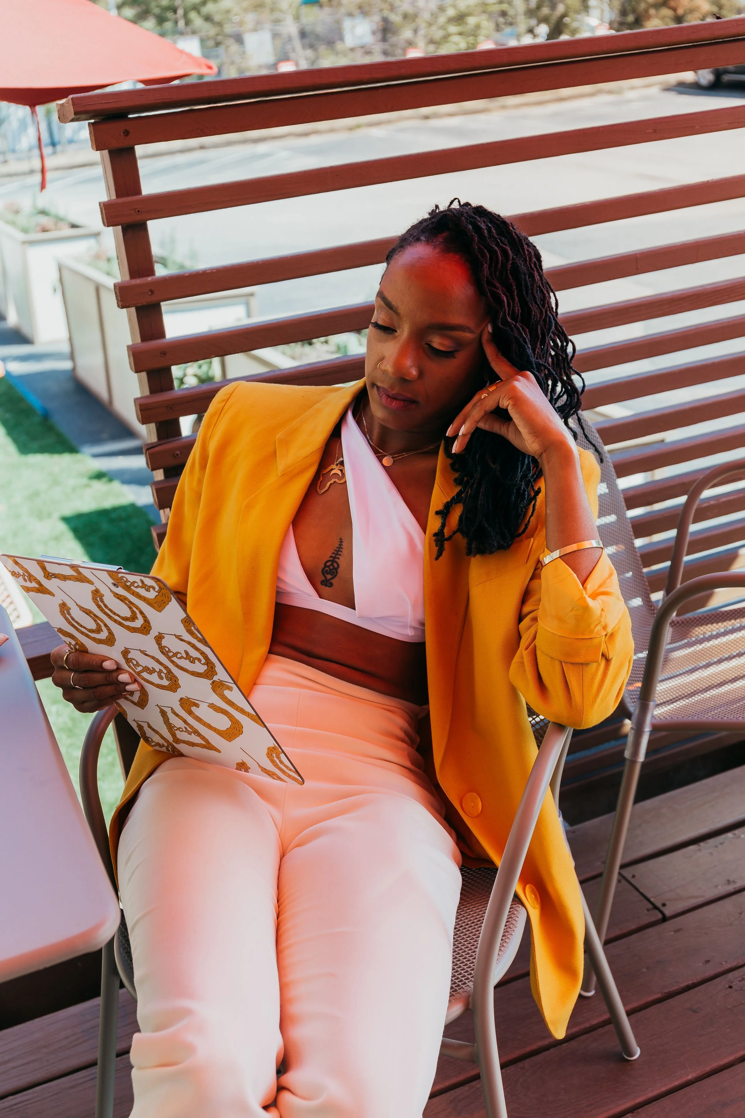 A woman sits on a chair outdoors, wearing a yellow blazer over a white and pink top, holding a patterned tablet and resting her head on her hand, appearing thoughtful.