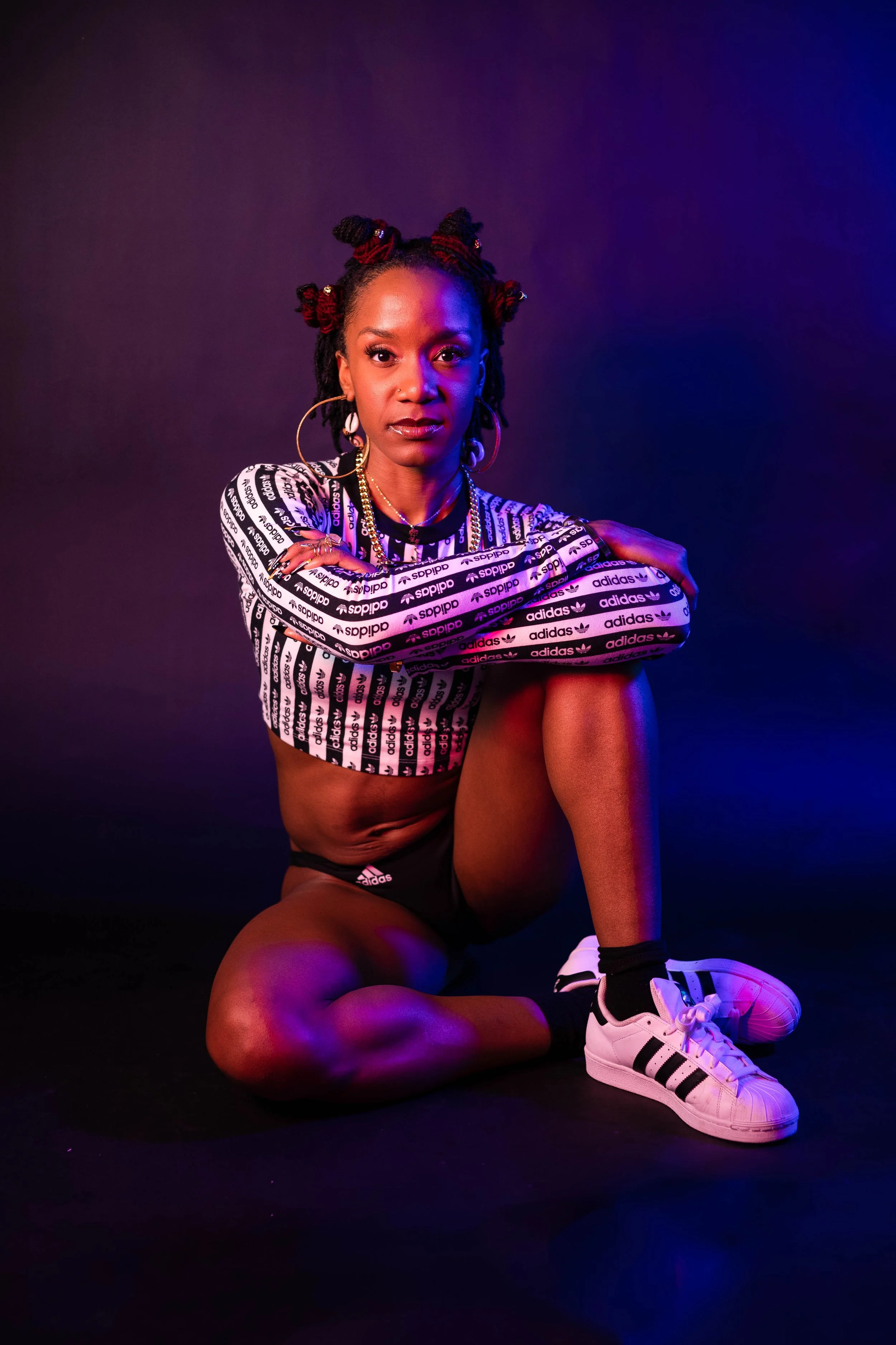A woman with styled hair in bantu knots, wearing Adidas sportswear, gold earrings and sneakers, sitting on the floor in a posed position with a colorful, vibrant lighting background.