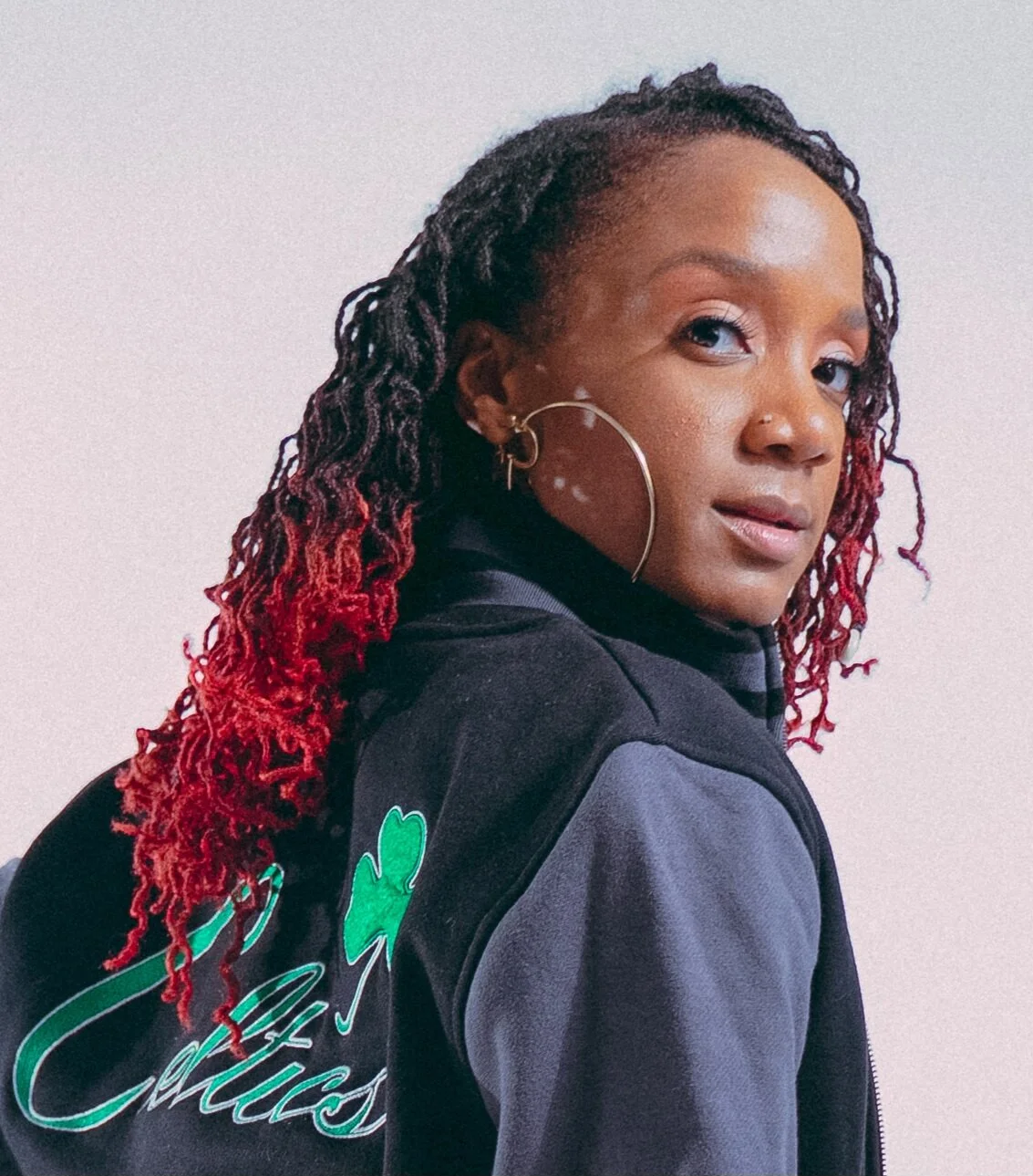 A woman with intricately styled dreadlocks, some dyed red, wearing large hoop earrings and a black and grey jacket with green shamrock design, standing against a plain white background.