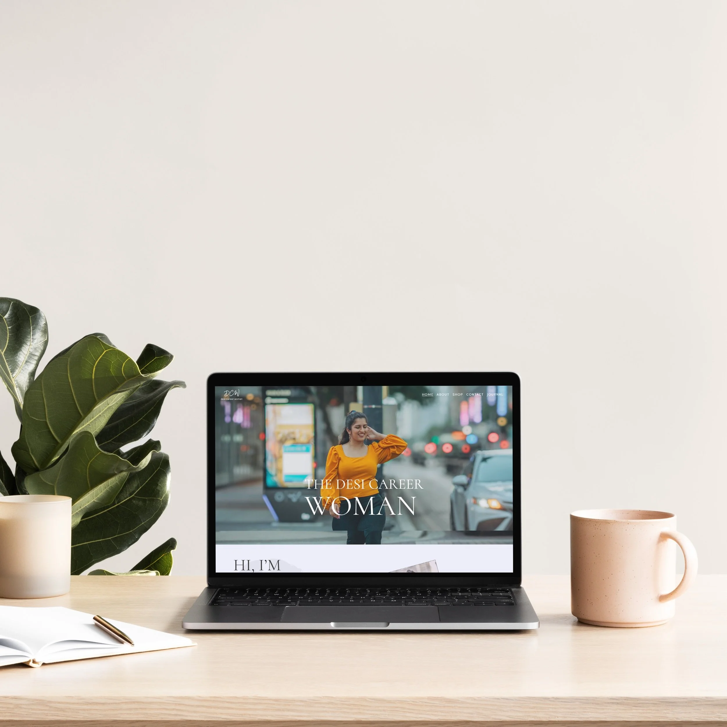 A desk with a laptop displaying a website titled 'The Desi Career Woman,' a plant in a white pot on the left, an open notebook with a pen, and a pink mug on the right.
