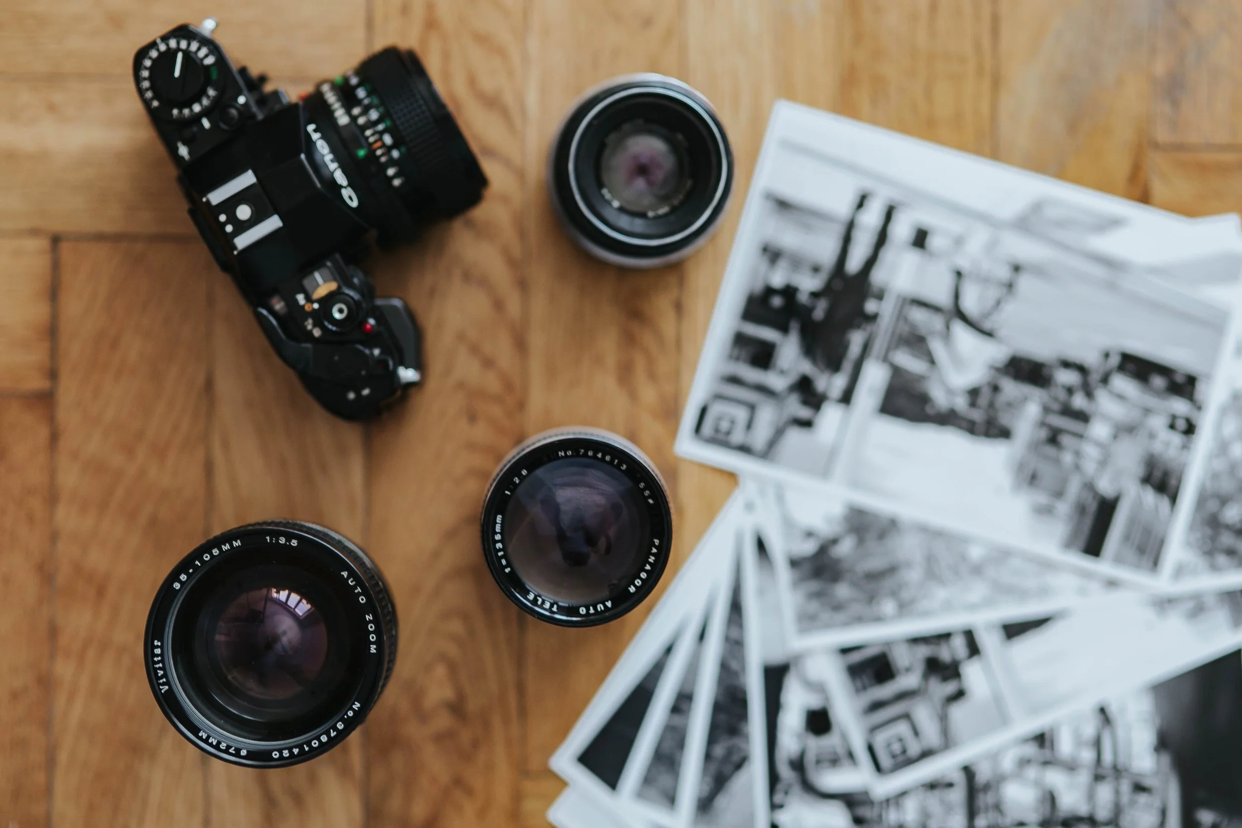 Camera, camera lenses, and black-and-white photographs on a wooden surface.