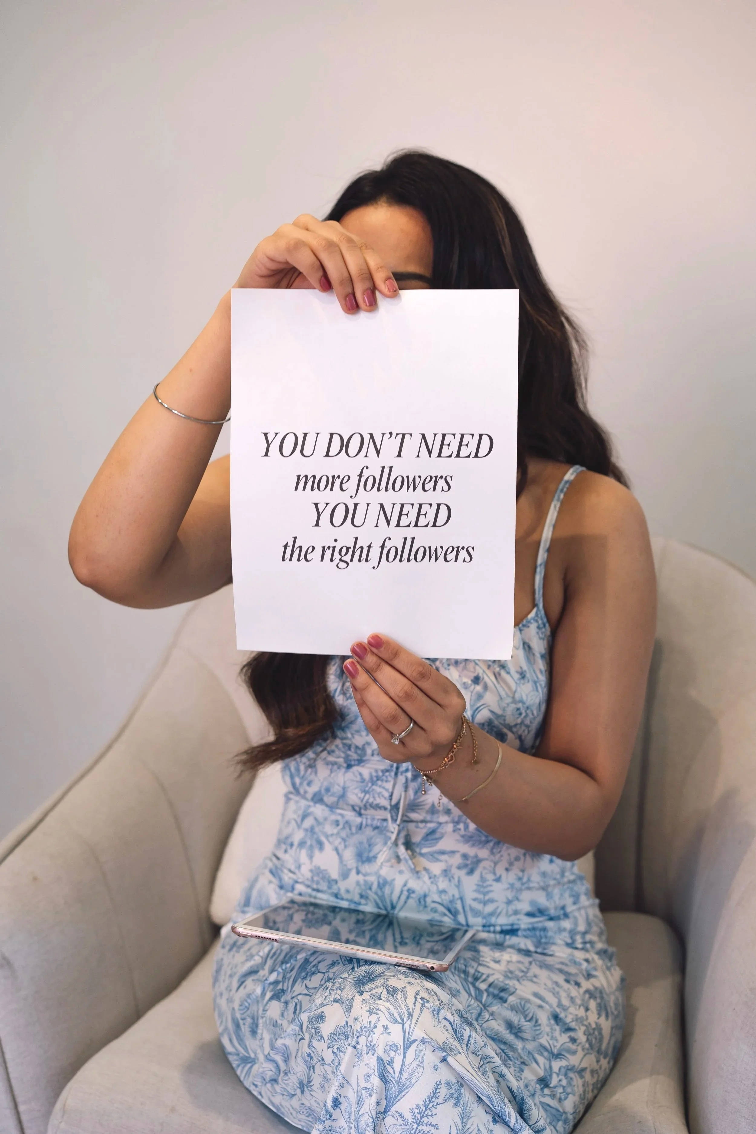 Woman sitting on a beige sofa holding a sign with a quote about followers covering her face. The quote says, 'You don't need more followers, you need the right followers.' The woman is wearing a floral dress, has dark hair, and is adorned with rings, bracelets, and a delicate necklace. A tablet rests on her lap.