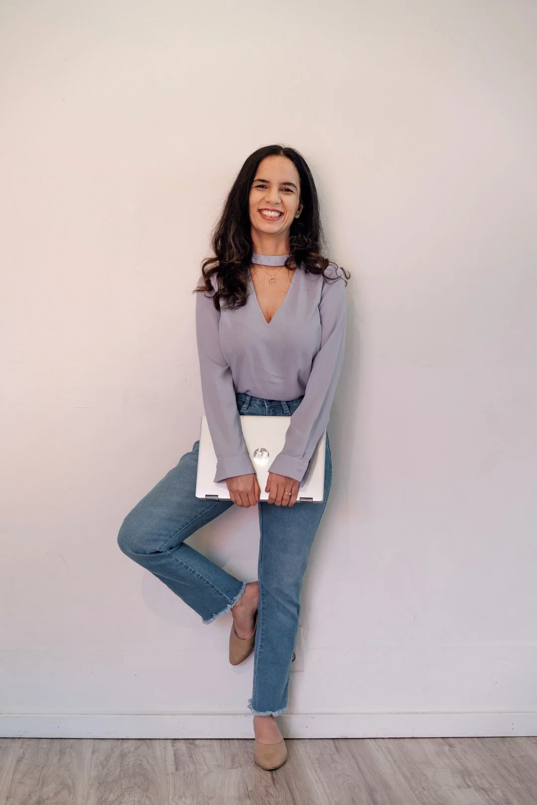 Young woman with long dark hair, wearing a light purple blouse, jeans, and beige shoes, standing against a plain white wall, holding a closed laptop.