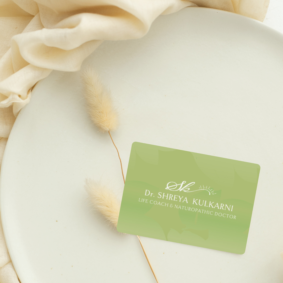 A business card for Dr. Shreya Kulkarni, a life coach and naturopathic doctor, resting on a light-colored surface surrounded by cream-colored fabric and a decorative fluffy plant stem.