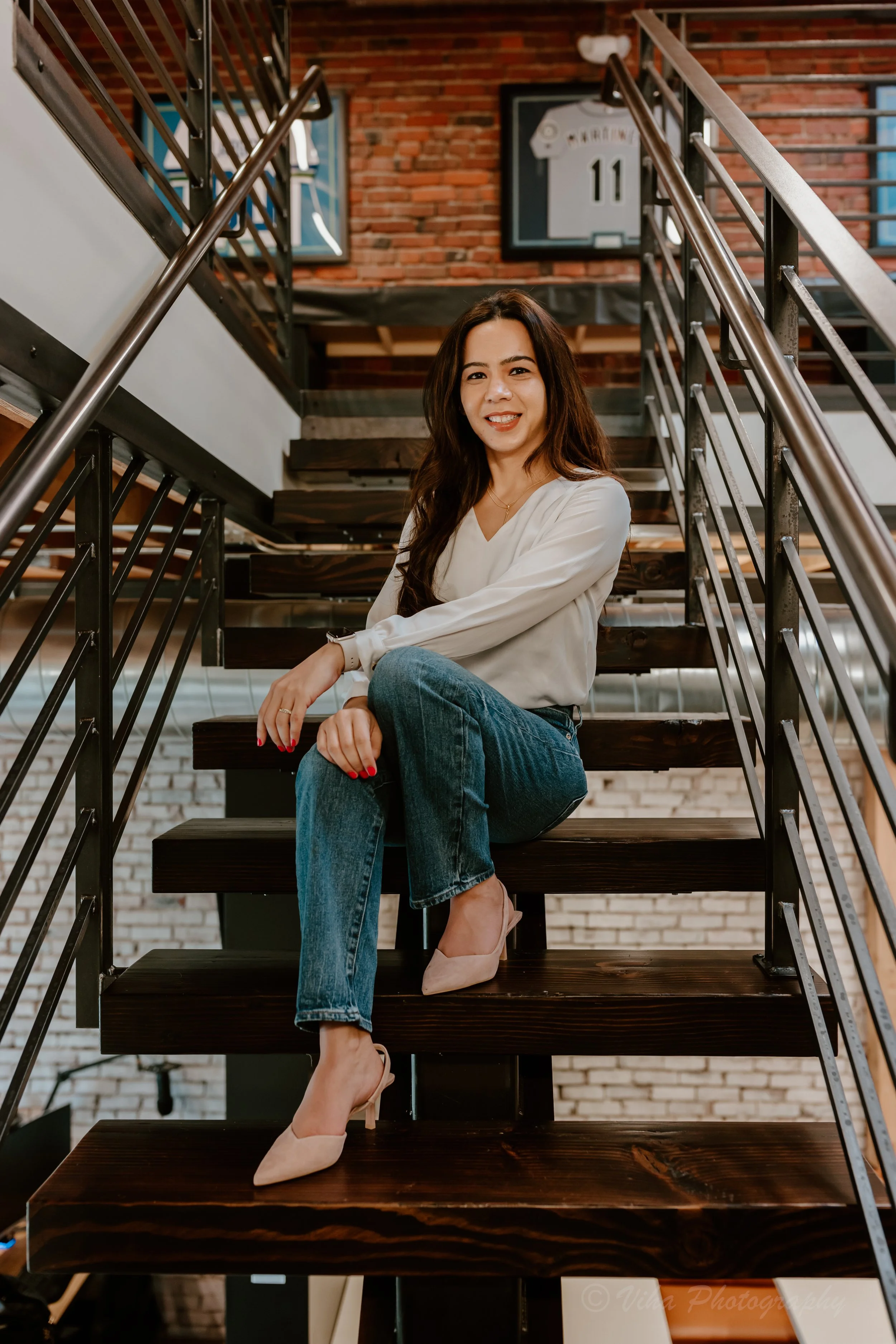 A woman with long dark hair, wearing a white blouse, blue jeans, and beige heels, sitting on dark wooden stairs inside a building with a brick wall background and sports jerseys framed on the wall.