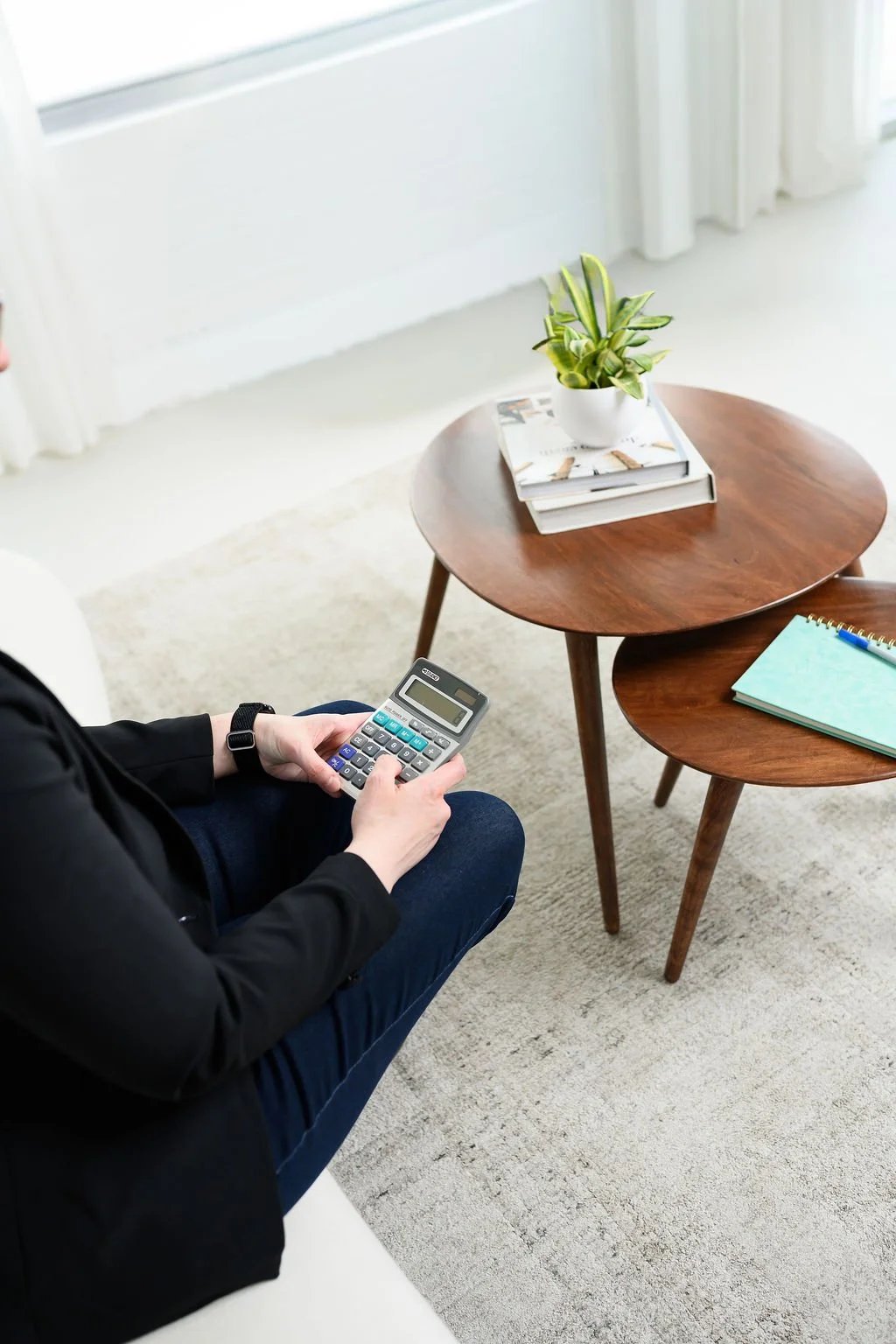 Person sitting on a white couch, holding a calculator, with two wooden coffee tables in front of them. One table has a plant in a white pot and a stack of magazines, the other has a notebook and pen. Light streaming through curtains.