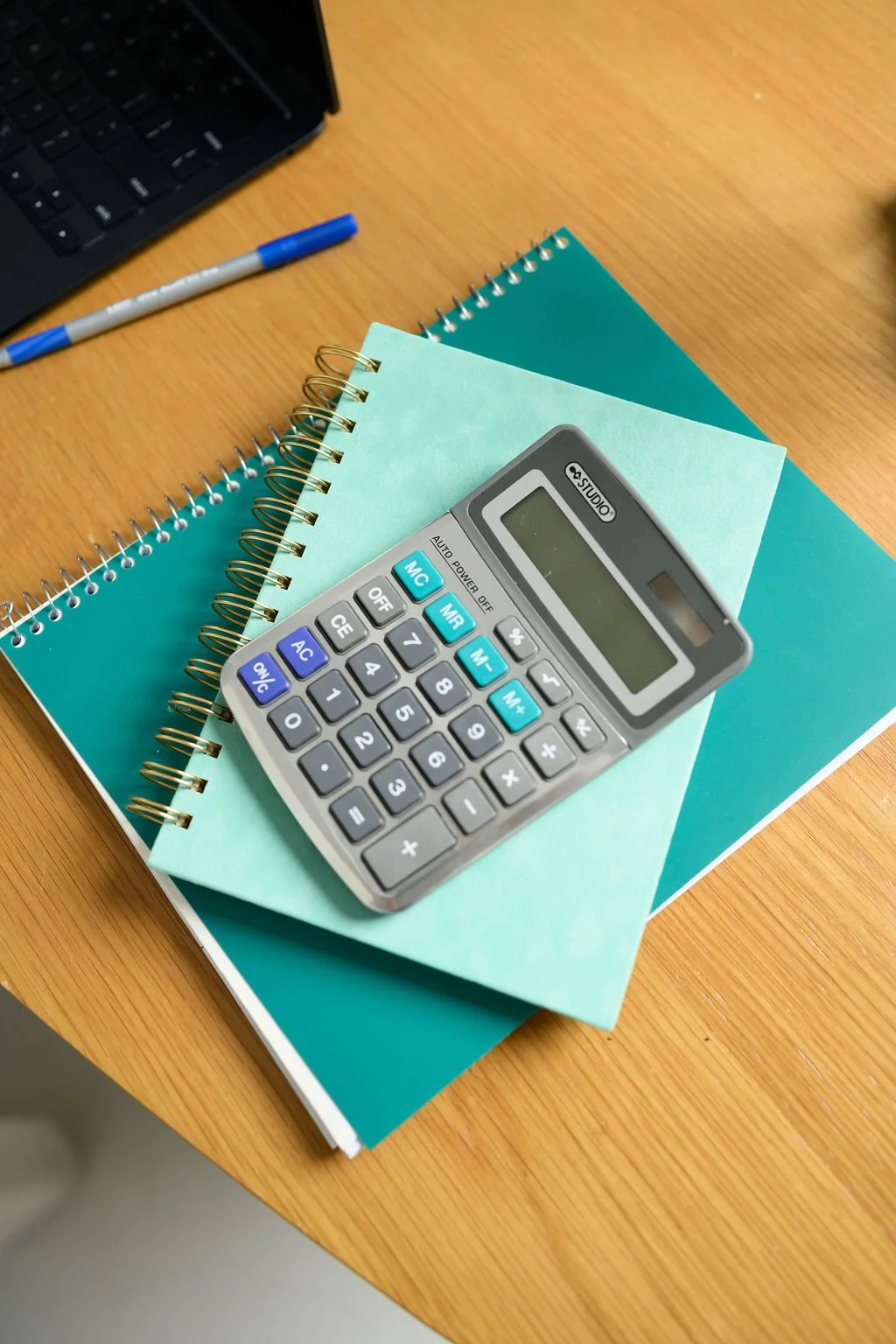A desk with two spiral-bound notebooks, a calculator, and a pen. The notebooks are teal and dark green. Part of a laptop is visible in the top corner.
