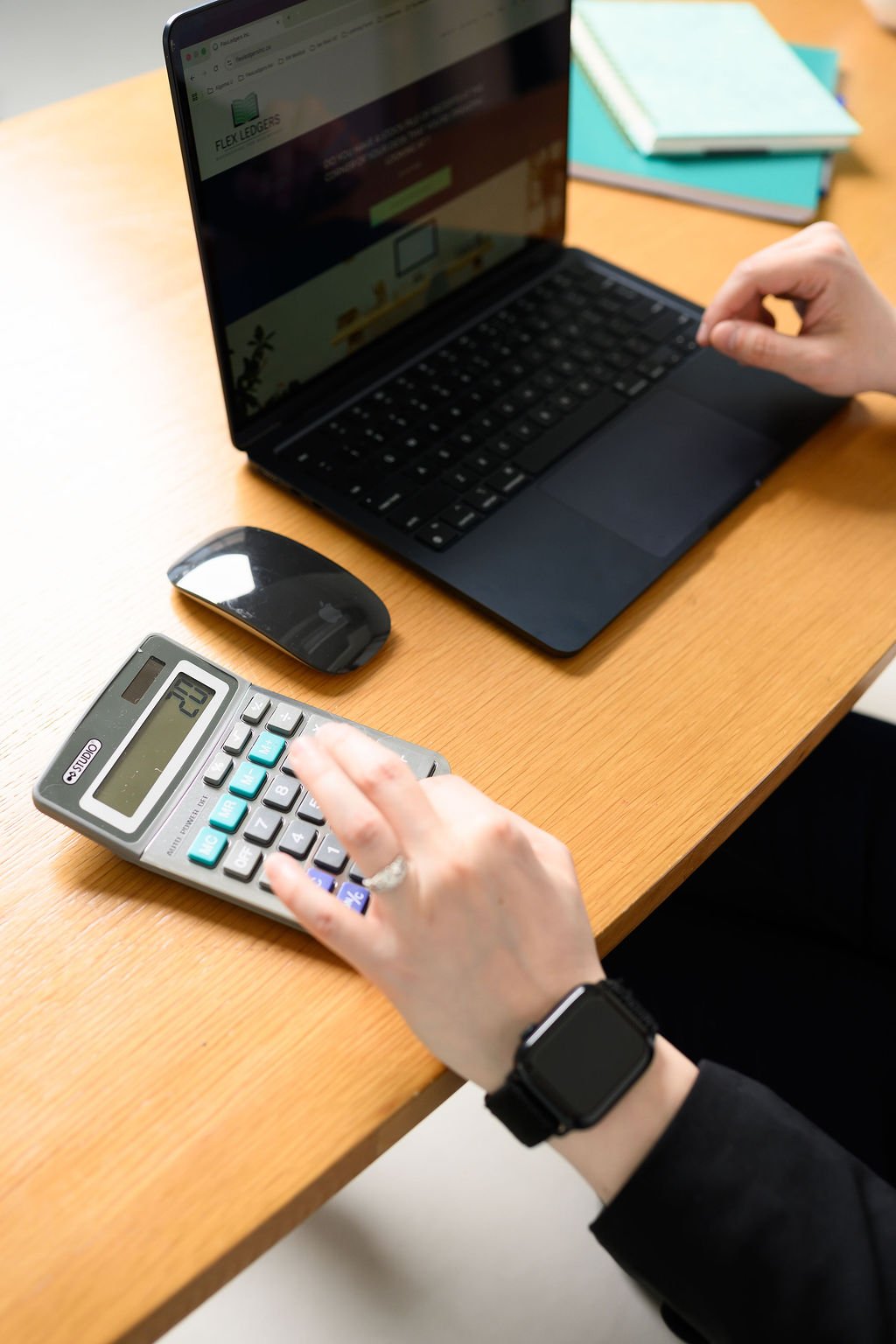 A person using a calculator and a laptop at a wooden desk, with a computer mouse nearby and notebooks in the background.