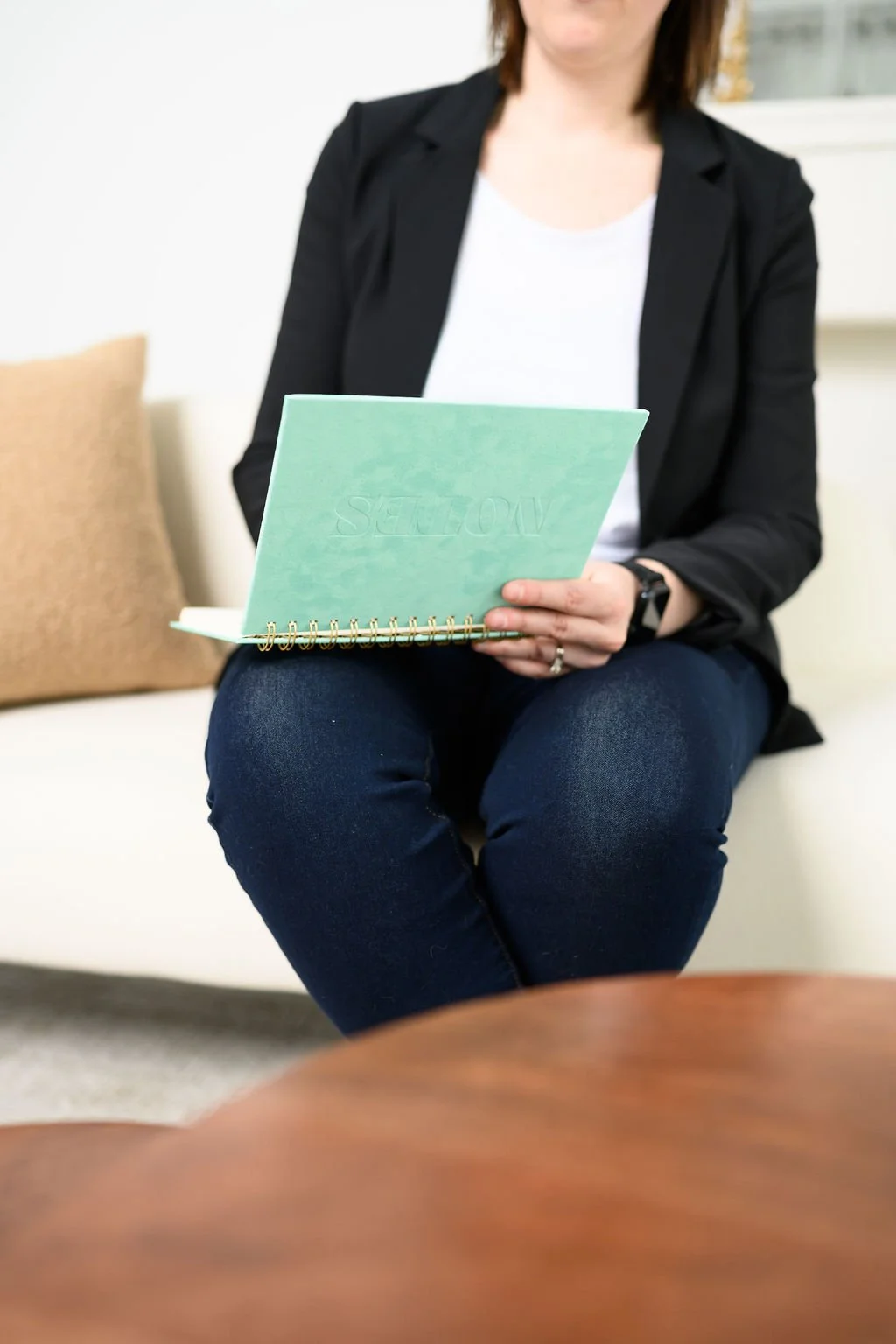 Woman sitting on a white couch holding a notebook with spiral binding, wearing a black blazer and white shirt, with a beige pillow and shelf in the background.