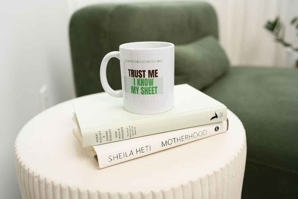 A white mug with the text "Trust Me I Know My Sheet" printed on it, placed on two stacked books on a small white table. The background includes a green couch and a potted plant.