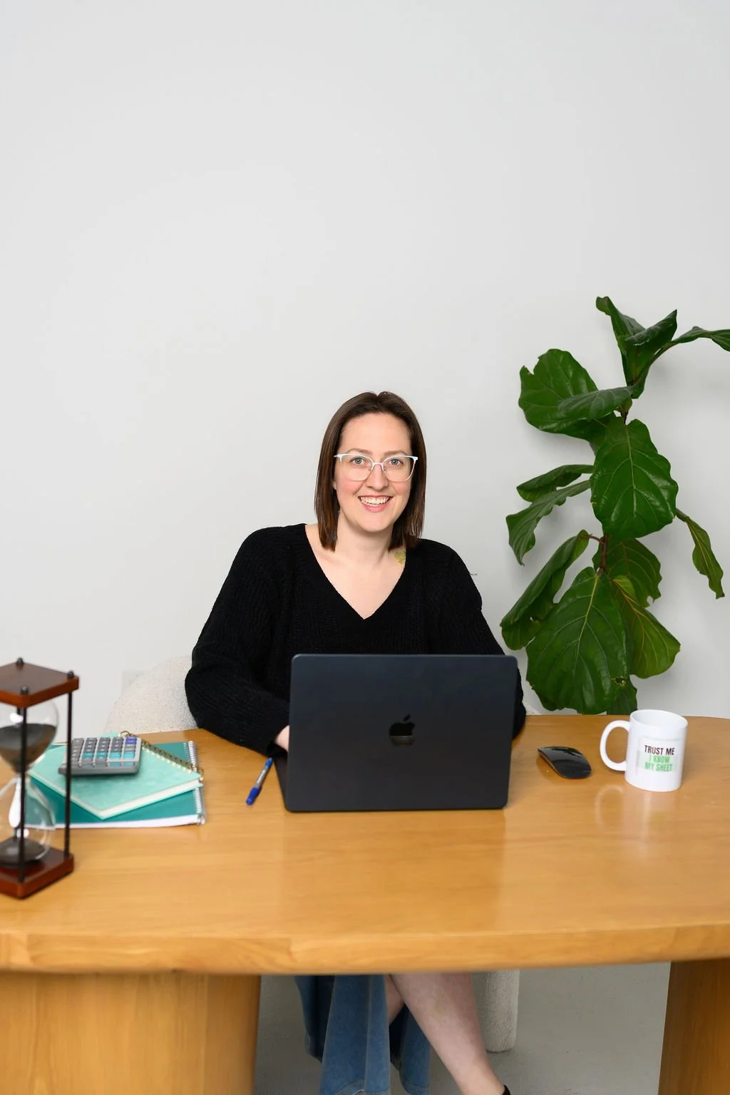 A woman sitting at a desk with a laptop, notebooks, a pen, a calculator, a coffee mug, and a mouse, smiling at the camera. There is a large green plant on the side and a small decor item on the desk.