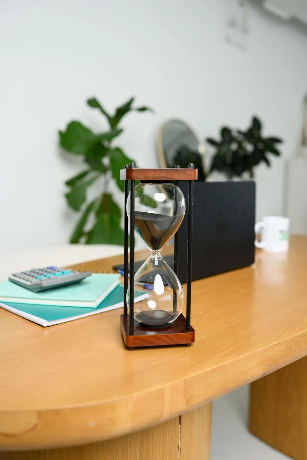 A wooden desk with a glass hourglass timer, some notebooks, a calculator, a white mug, and a black rectangular object, with two large green potted plants in the background.
