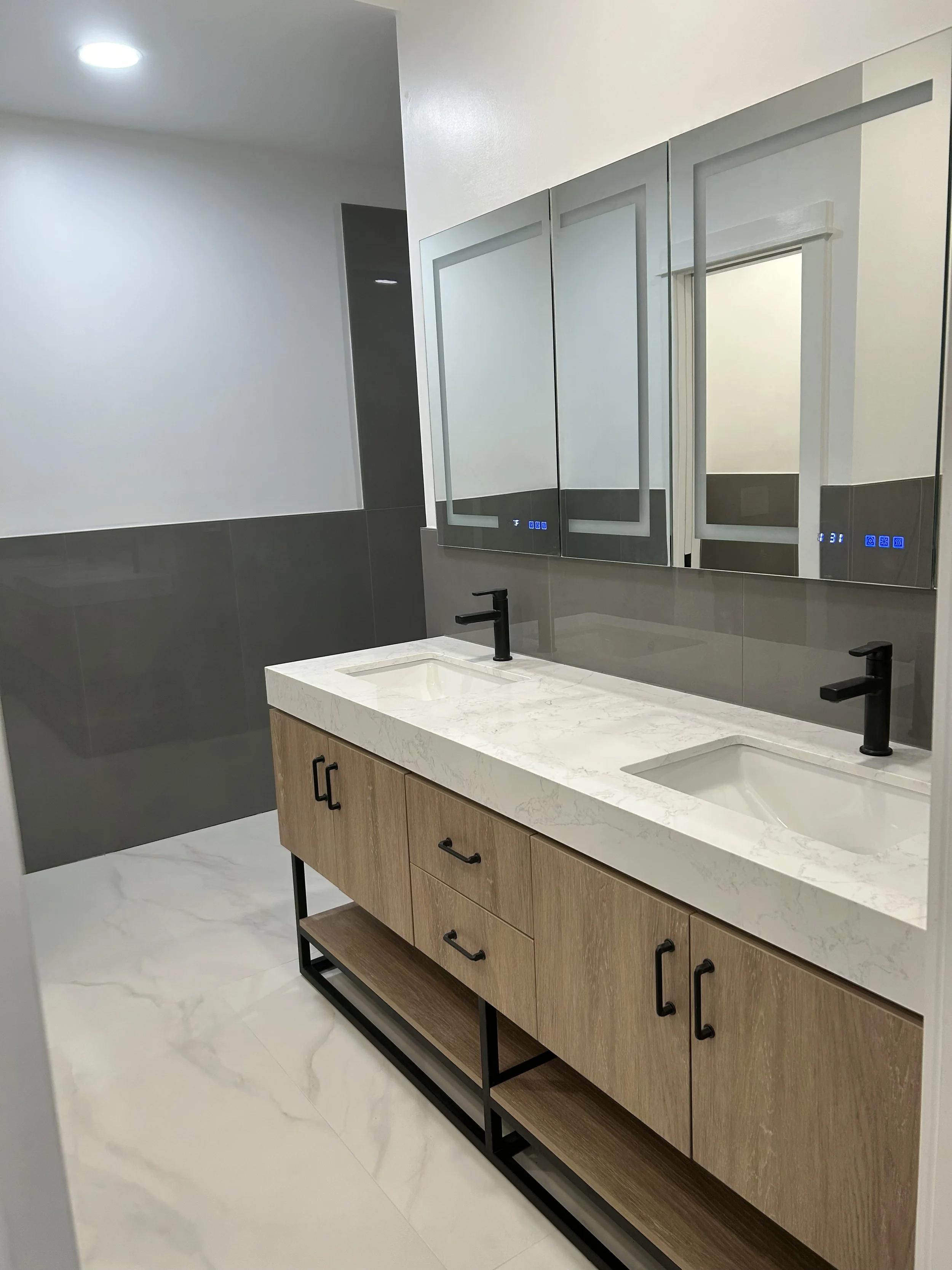 Modern bathroom with dual sinks, black faucets, wooden vanity with open shelf, large mirror with touch controls, and gray and white tile design.
