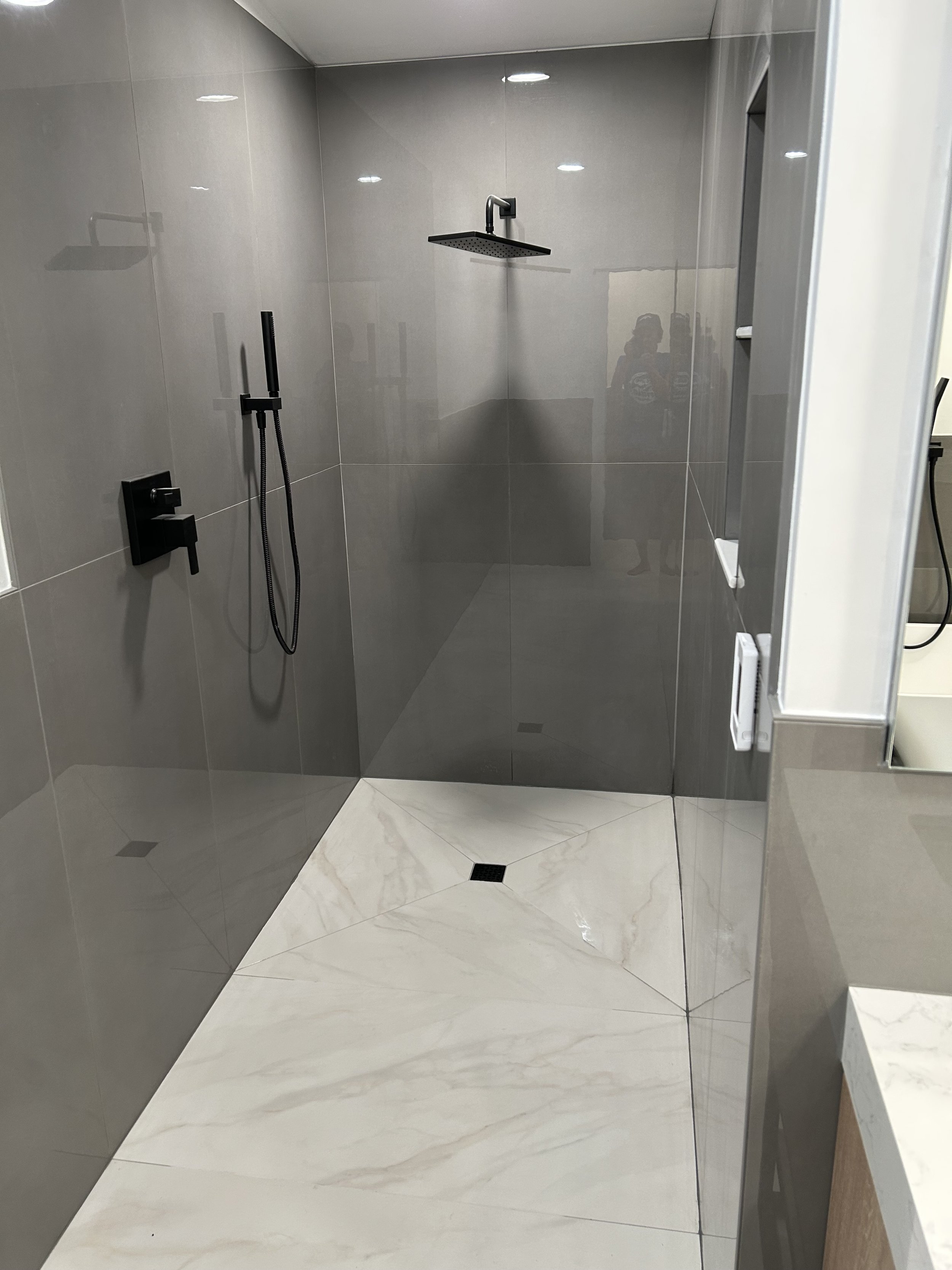 Modern walk-in shower with a black rainfall showerhead, a black handheld shower, gray tiled walls, and beige marble-like floor tiles.