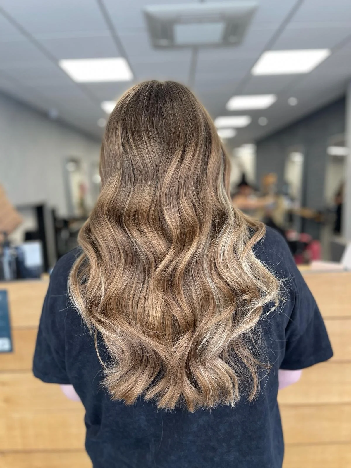 Back view of a woman with long, wavy, light brown hair with blonde highlights inside a hair salon.