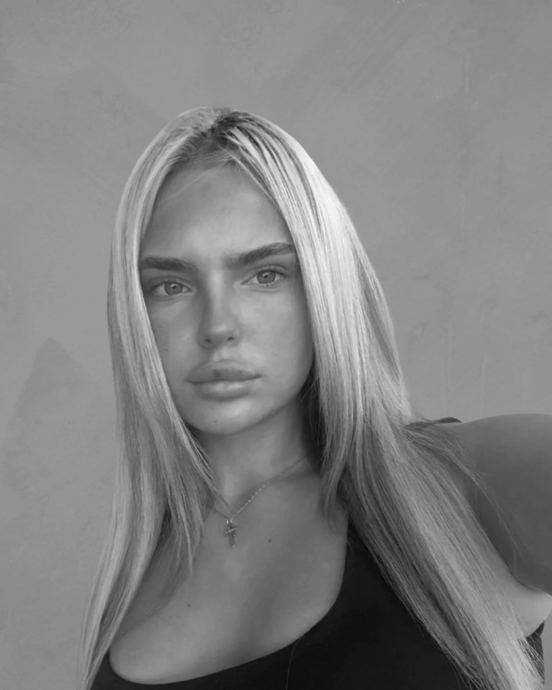 A black and white portrait of a young woman with long, straight blonde hair, wearing a dark top and a necklace with a cross pendant.