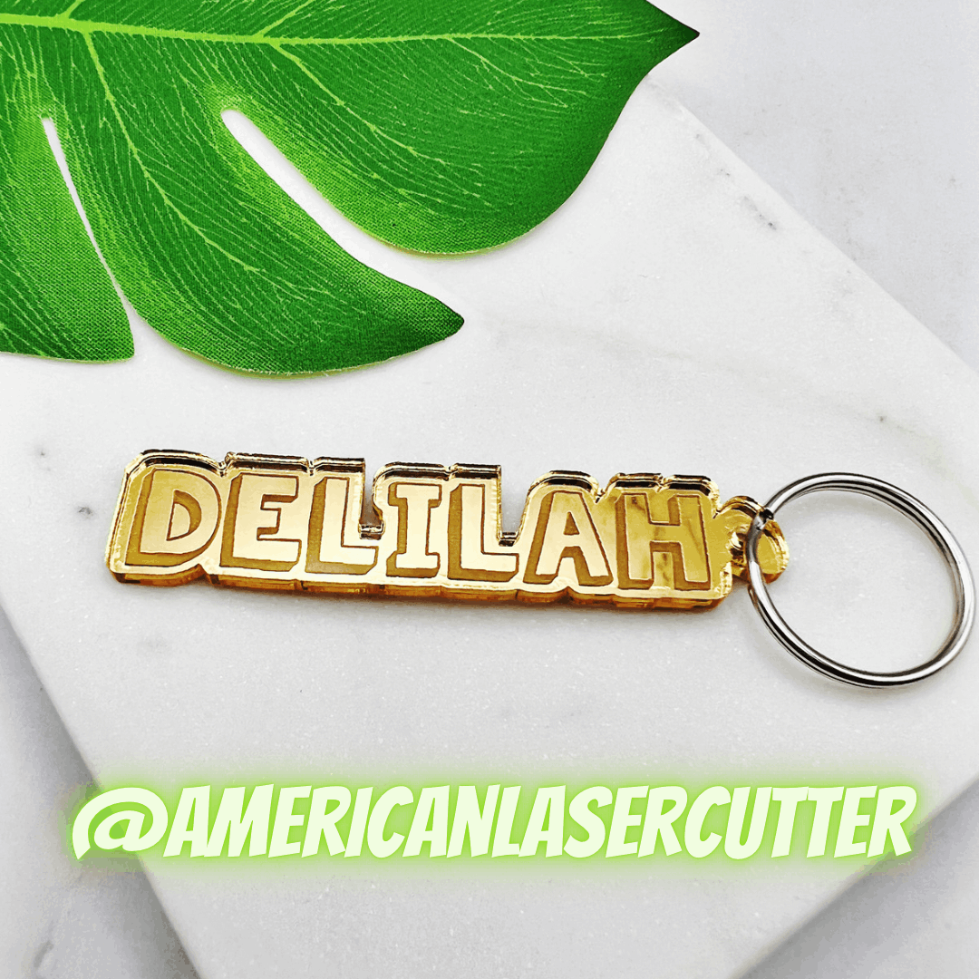 Gold-colored keychain with the name 'DELLILAH' in block letters, attached to a keyring, placed on a white surface with a green leaf in the background and @AMERICANLASERCUTTER written in neon green text.