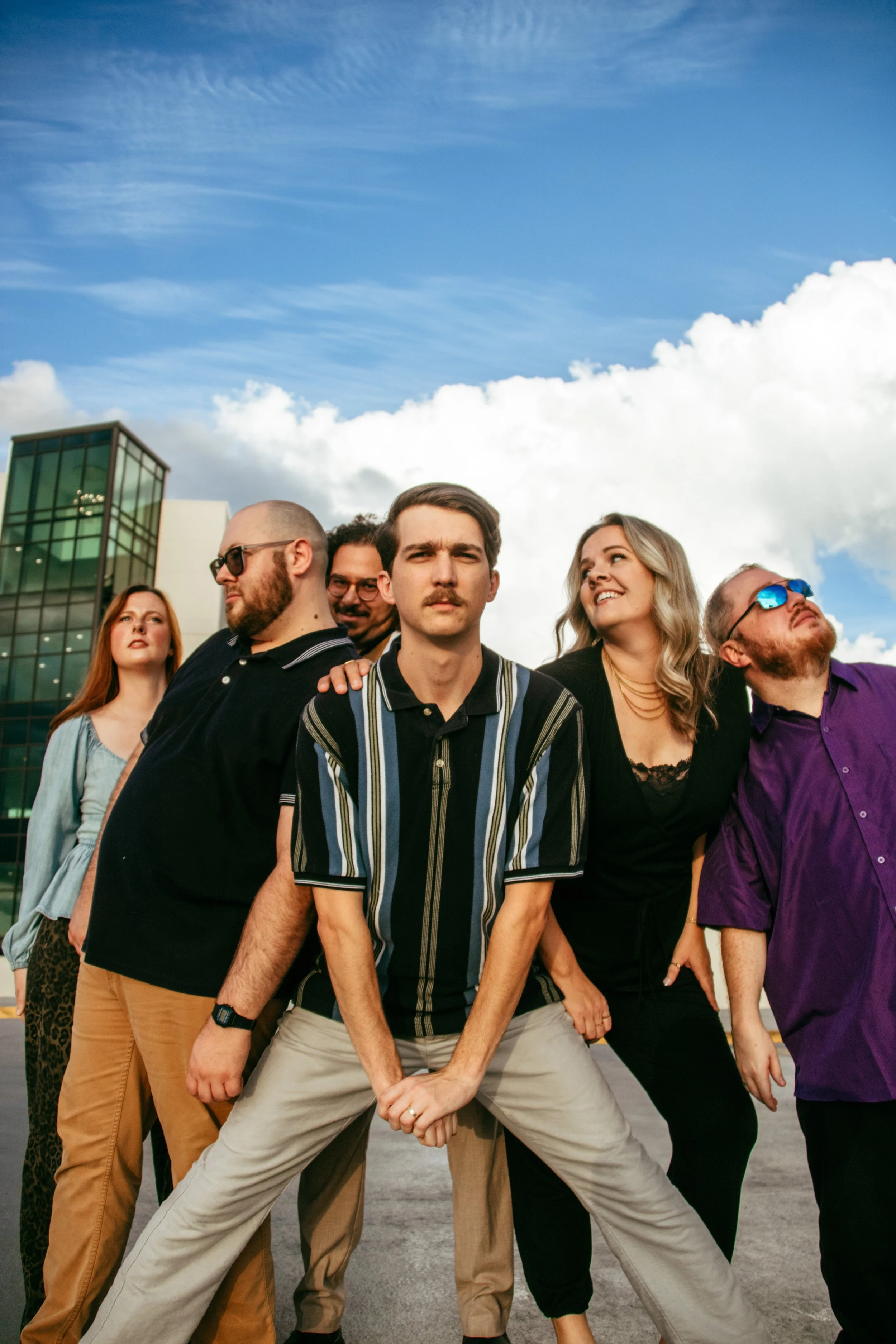 Group of six diverse young adults posing outdoors on a city street with a modern glass building and a partly cloudy sky in the background.