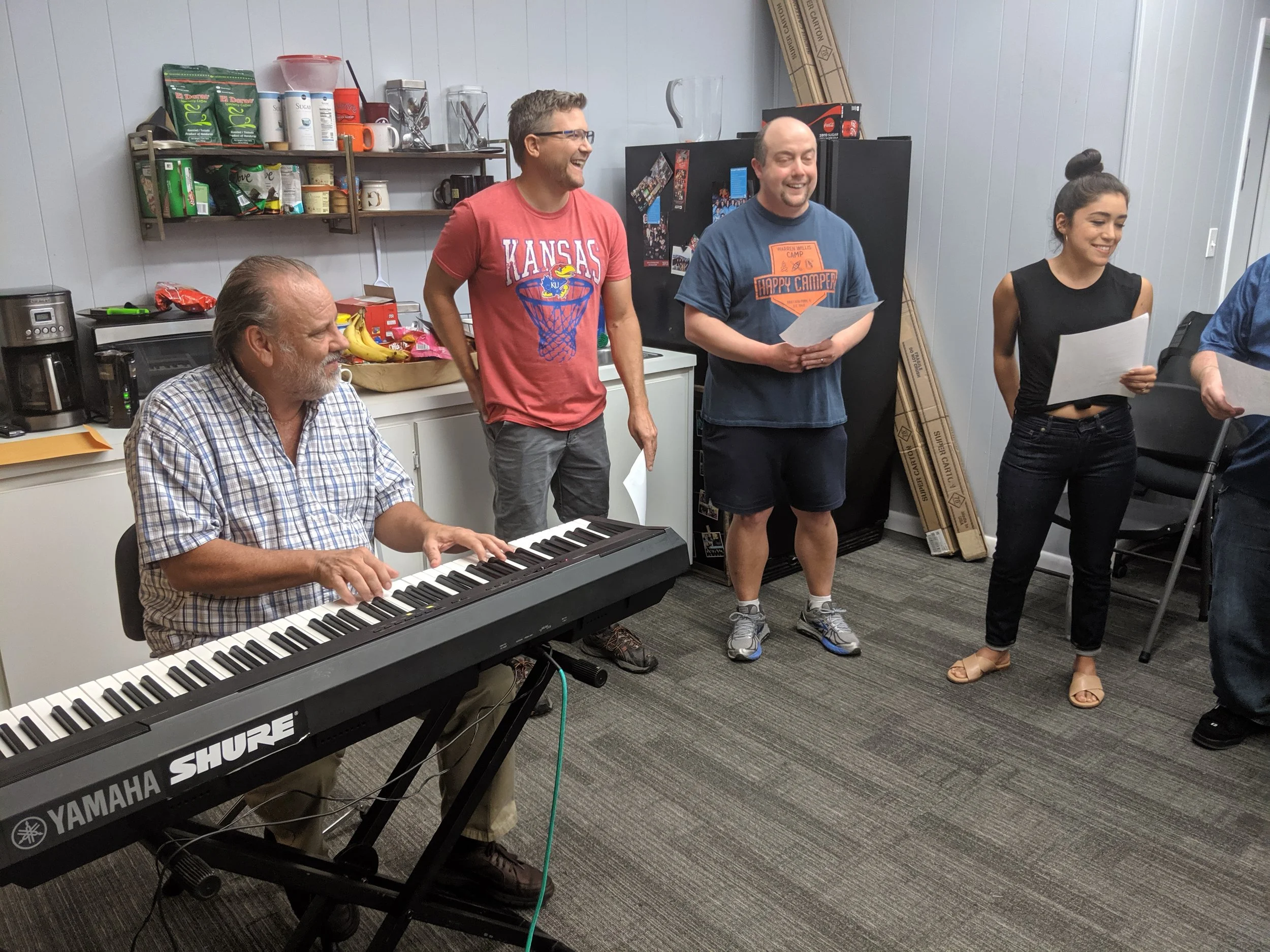 Group of five people singing and playing piano in a room with a gray wall and kitchen supplies in the background.