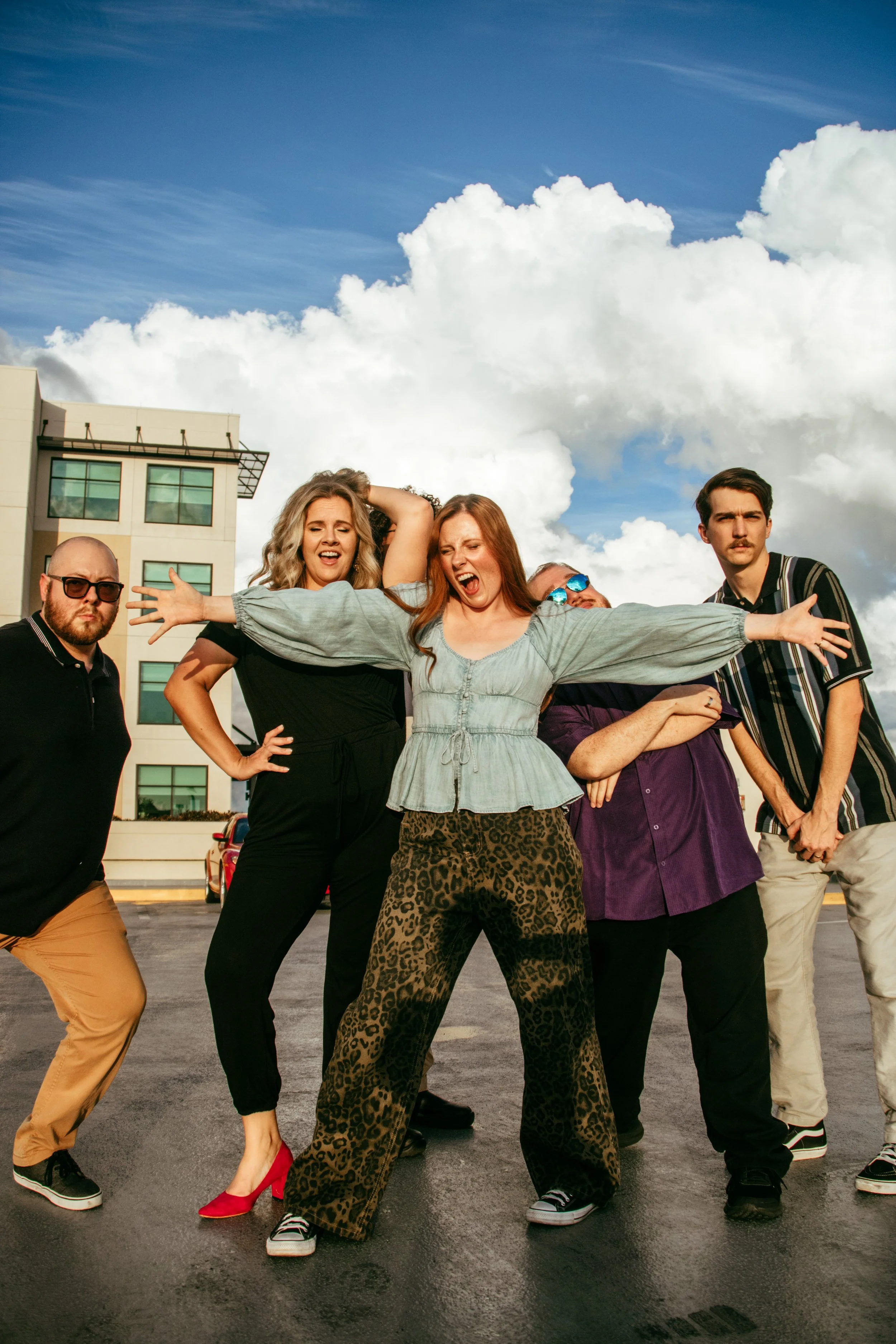 Group of five young adults posing playfully outdoors on a cloudy day, with a modern building in the background.