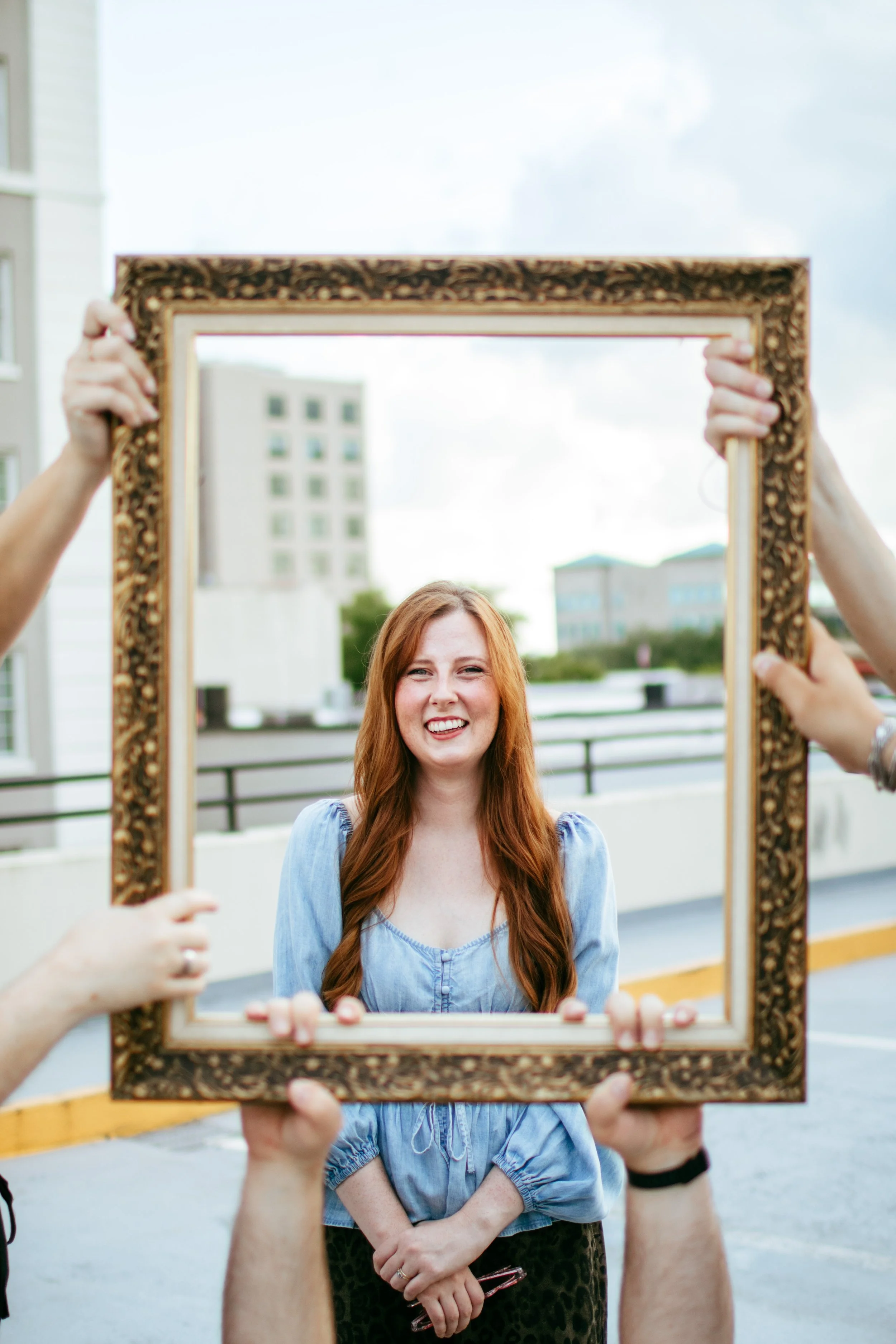 A woman with long red hair, wearing a blue shirt, standing outdoors and smiling as her picture is taken through an ornate empty picture frame held by four people.