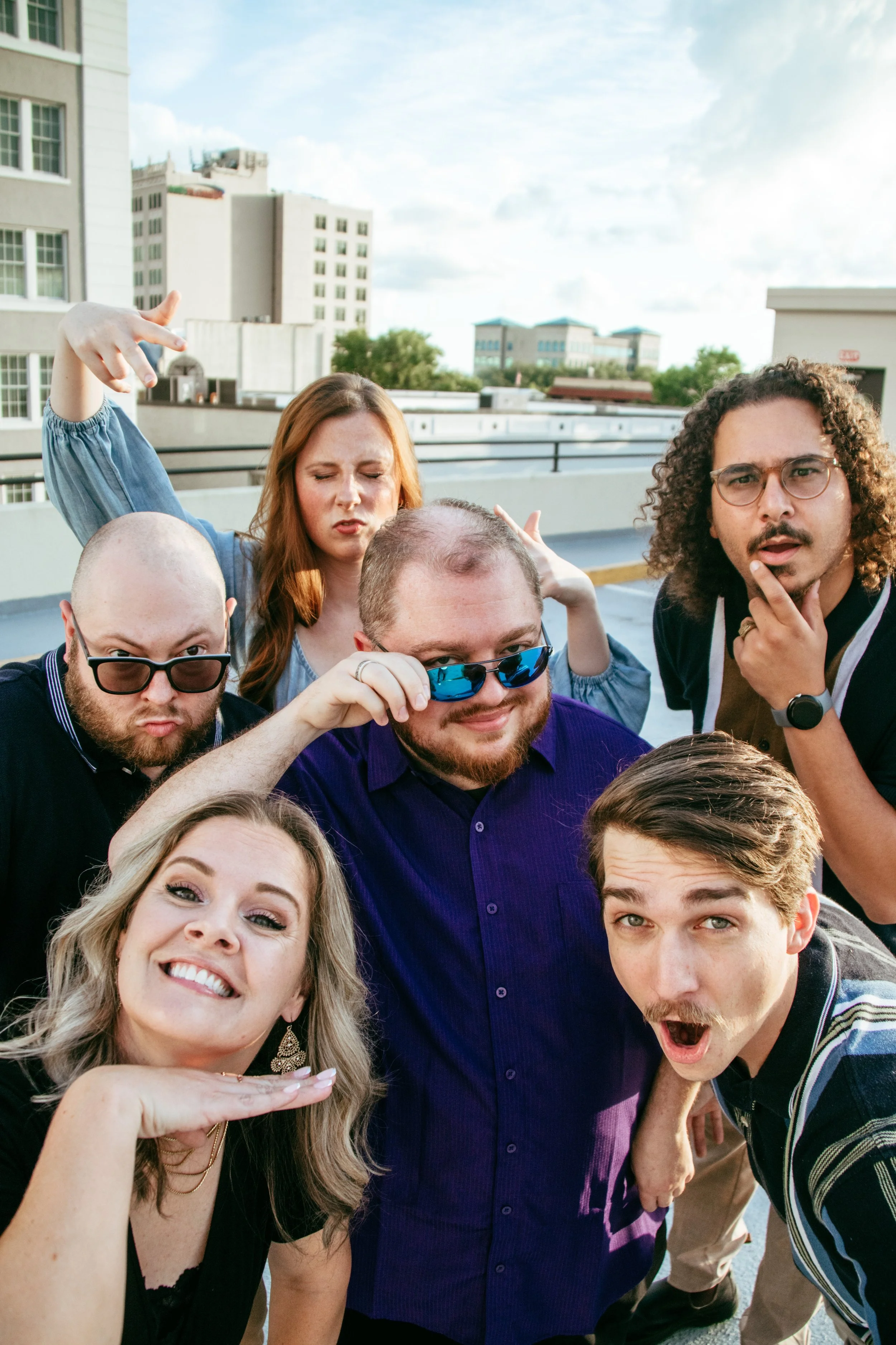 Group of six young adults having fun on rooftop terrace, posing with playful expressions, some wearing sunglasses, cityscape in background.